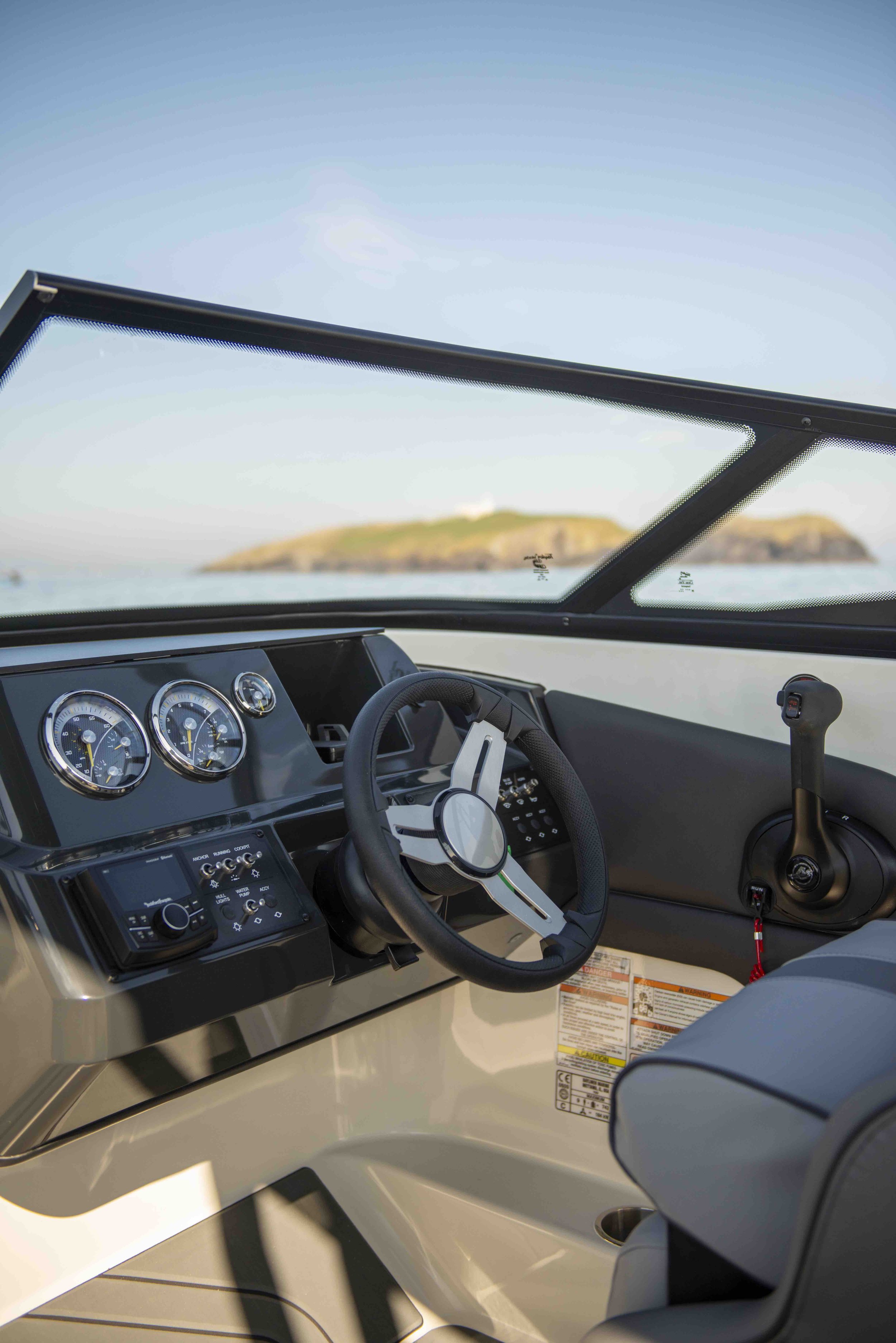 Boat cockpit interior with steering wheel and control panel under clear blue sky, distant land visible through windshield.