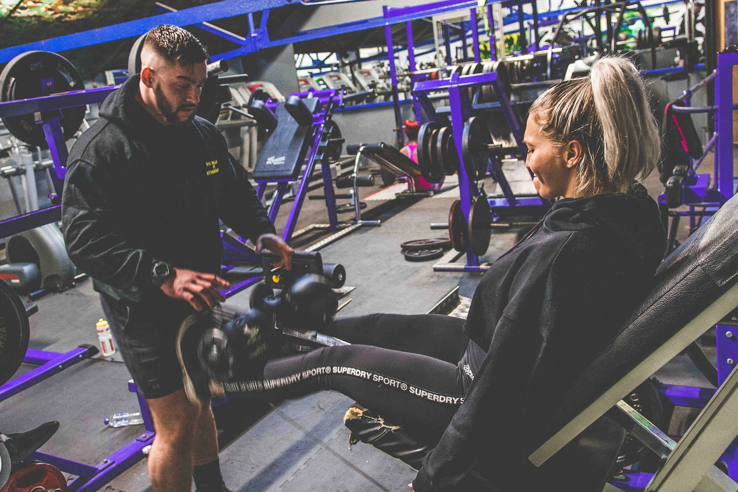 Woman in black workout clothes seated on a leg press machine, with a man in black hoodie assisting her in a gym setting.