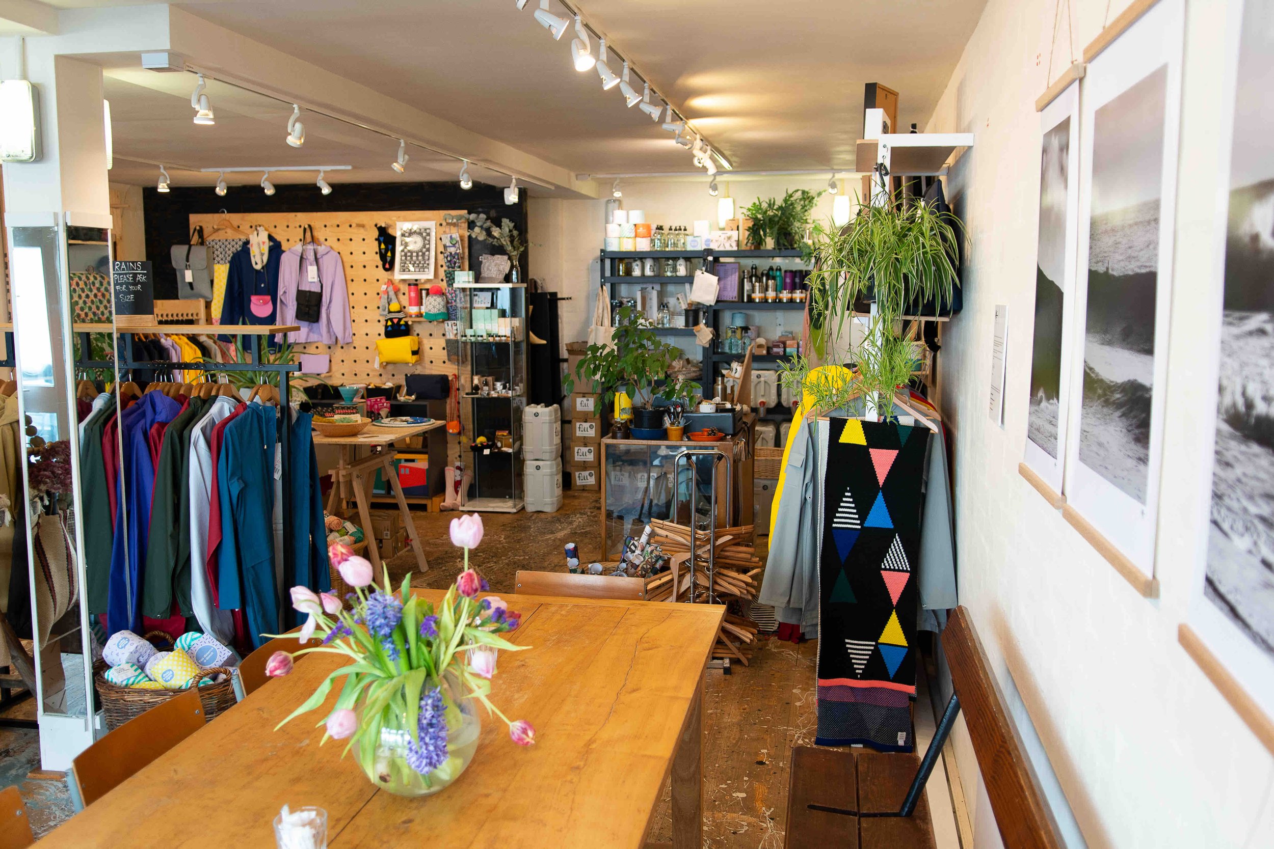 Interior of a small boutique store with colorful clothing on racks, a wooden table with a vase of flowers, assorted plants, and various items on display shelves.