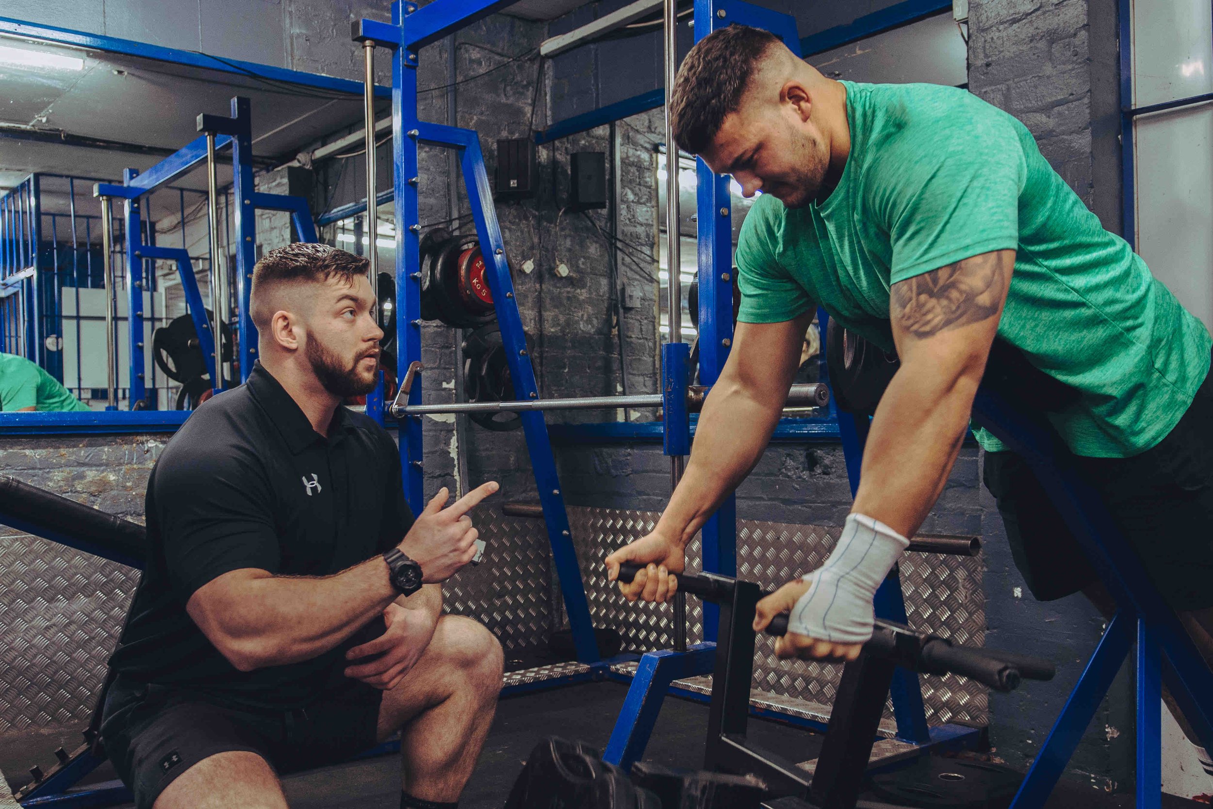 Two men in a gym, one wearing a black shirt is squatting and gesturing, and the other in a green shirt is using a weight machine, with wrist support, in a fitness environment.