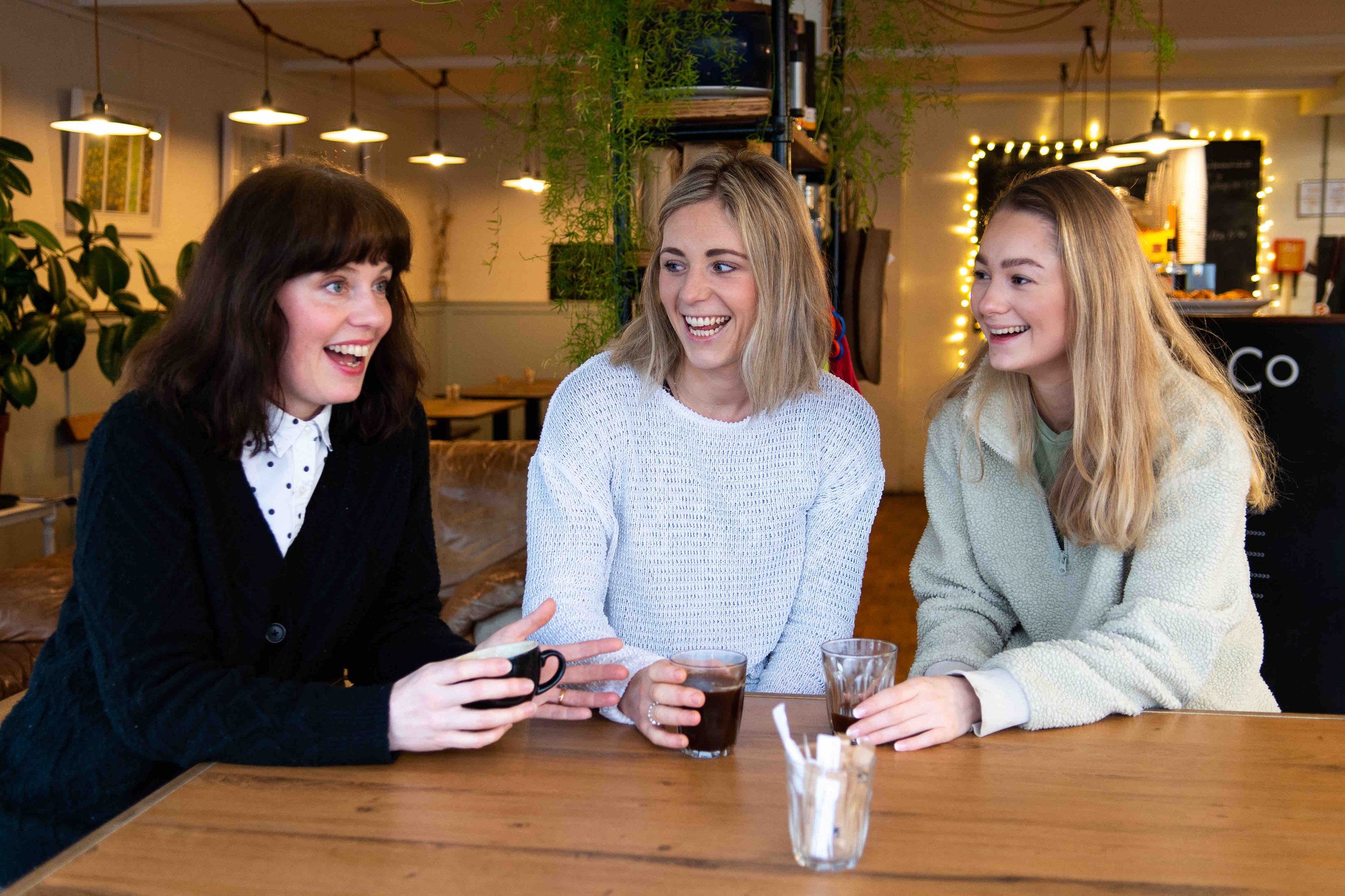 Three women sitting at a café table, laughing and holding drinks.