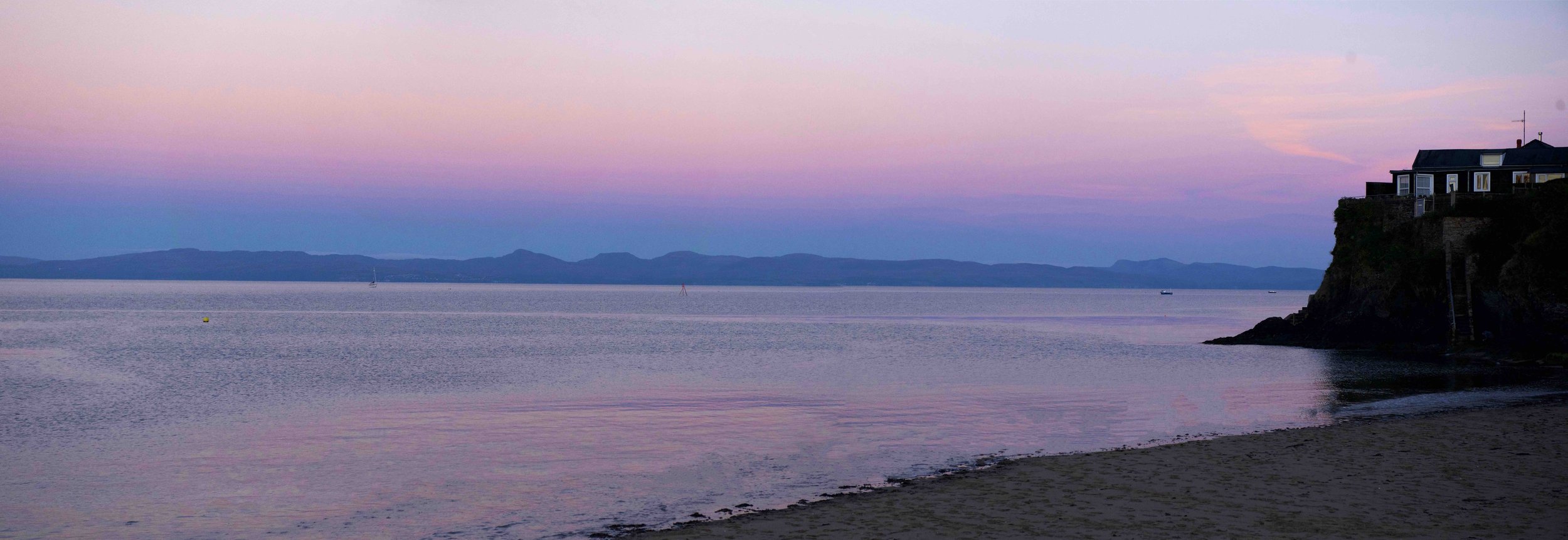 A serene coastal scene at dusk with a sandy beach in the foreground, calm water, distant hills, and a building situated on a cliff to the right. The sky displays soft pastel shades of pink and purple.