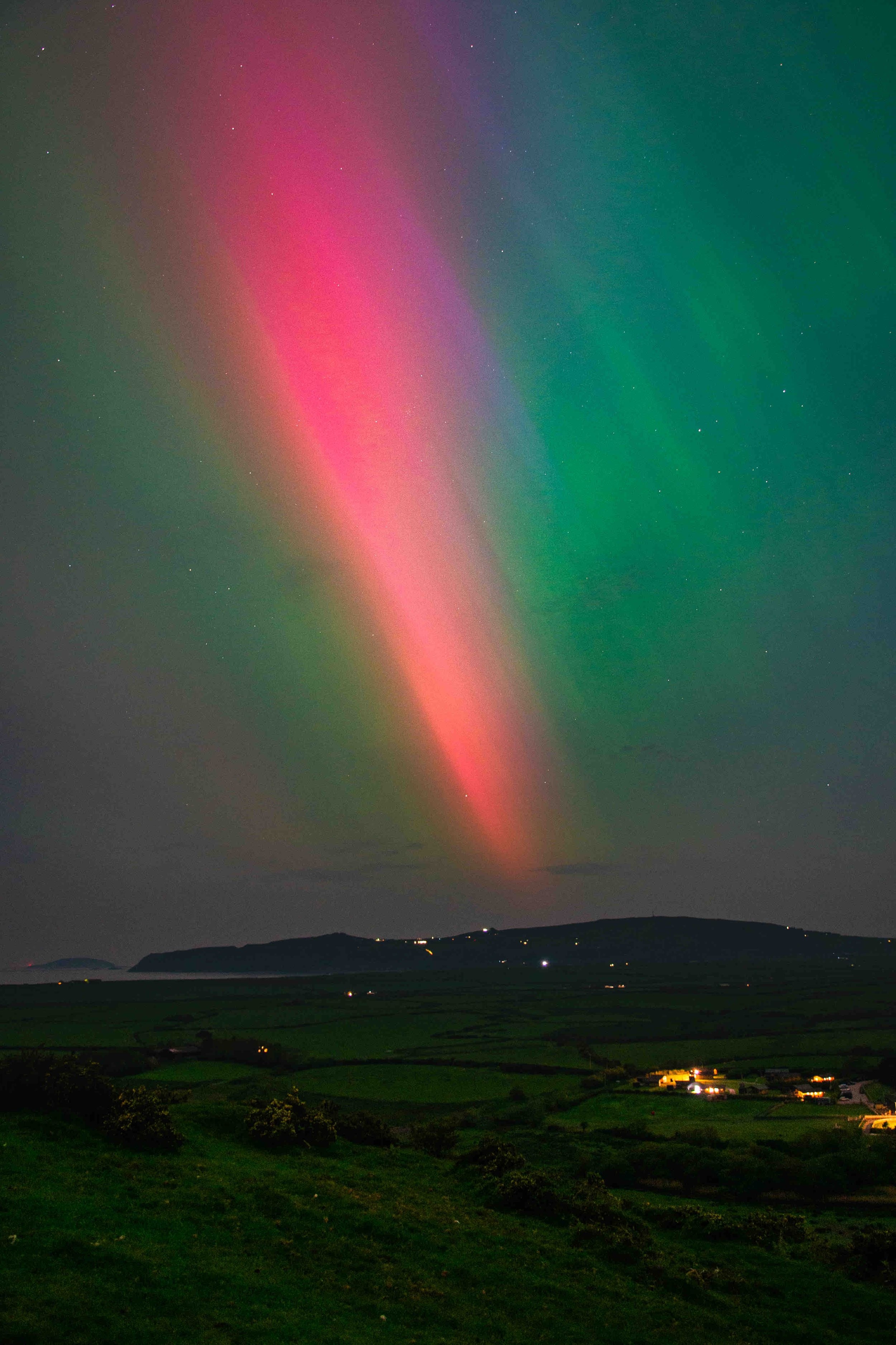 Colorful aurora in the night sky over a dark landscape with faint lights from buildings and hills in the distance.