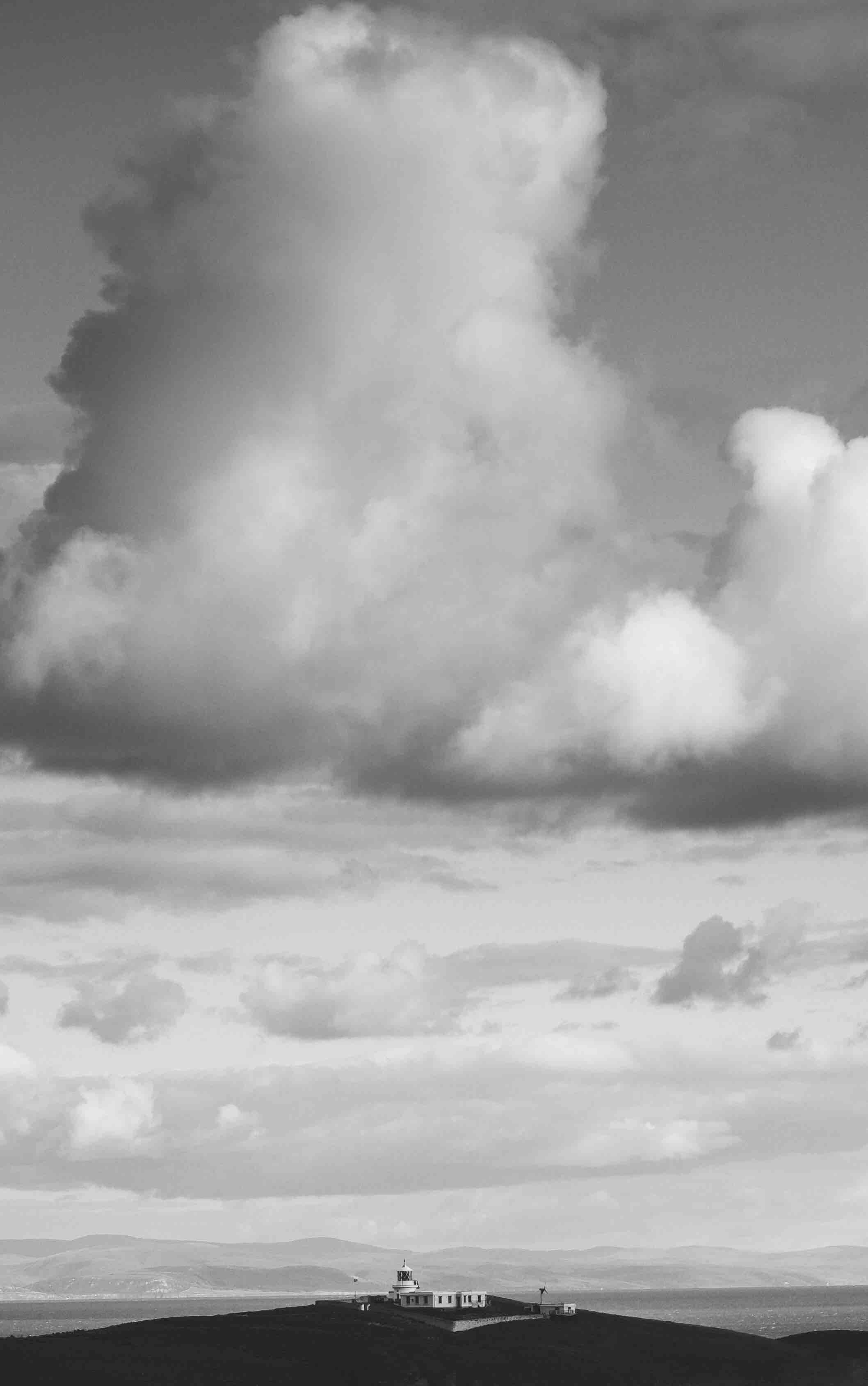 Black and white photo of a cape with a lighthouse, large clouds above, and distant hills in the background.