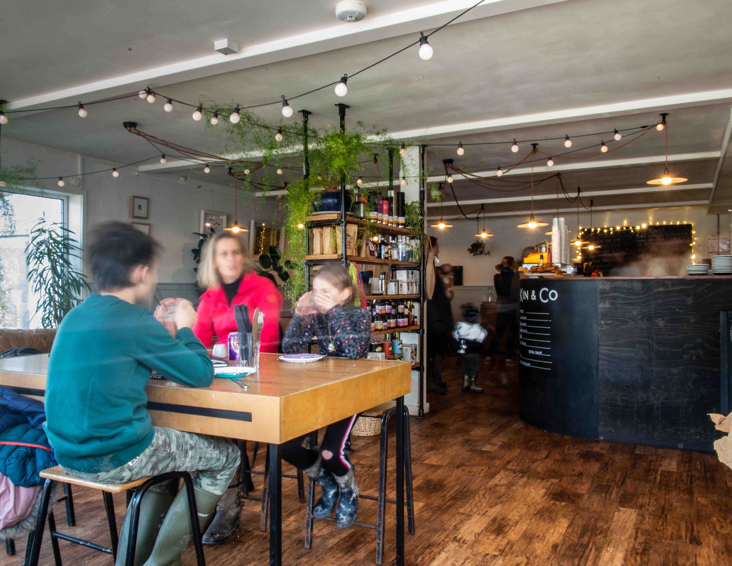 Cozy cafe interior with people sitting at a wooden table, surrounded by hanging lights, plants, and a black counter. Shelves with various products are visible in the background.