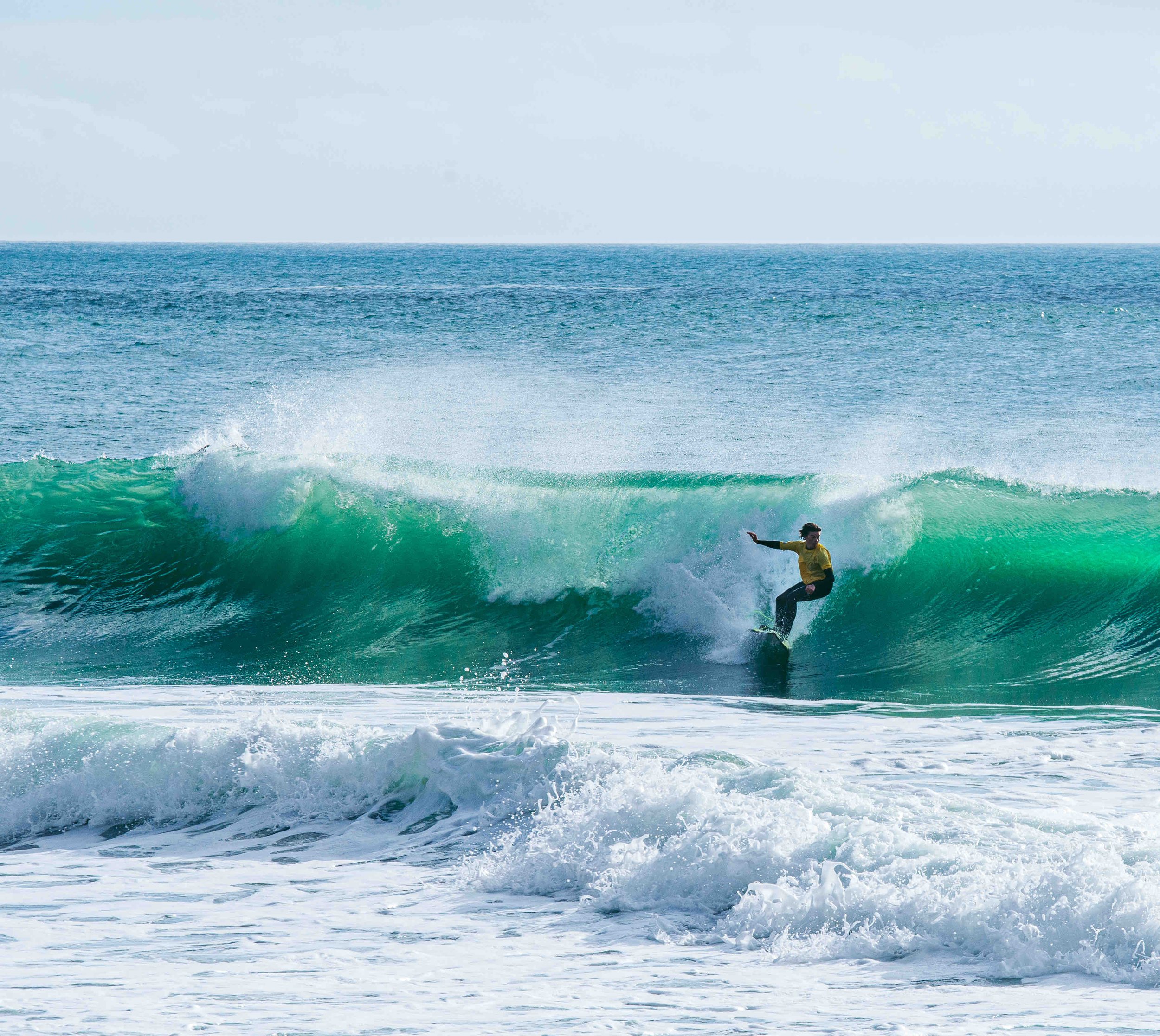 Surfer riding a green wave in the ocean, wearing a yellow shirt and black wetsuit, clear sky in the background.