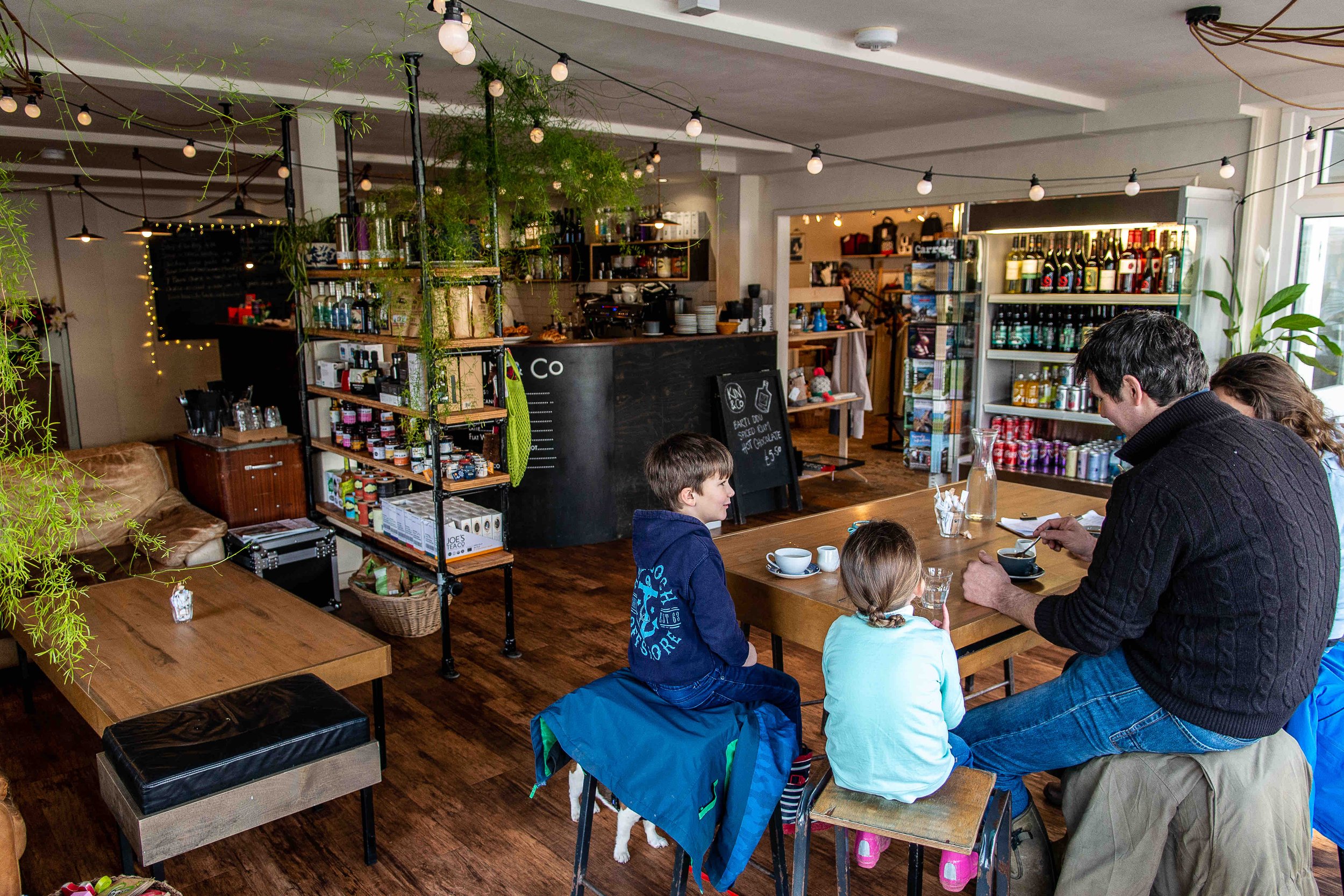 Cozy café interior with people sitting at a wooden table, surrounded by shelves of goods and a bar area. Decorative lights and plants are hanging from the ceiling. Shelves display various drinks and products. Chalkboard with menu near the counter.