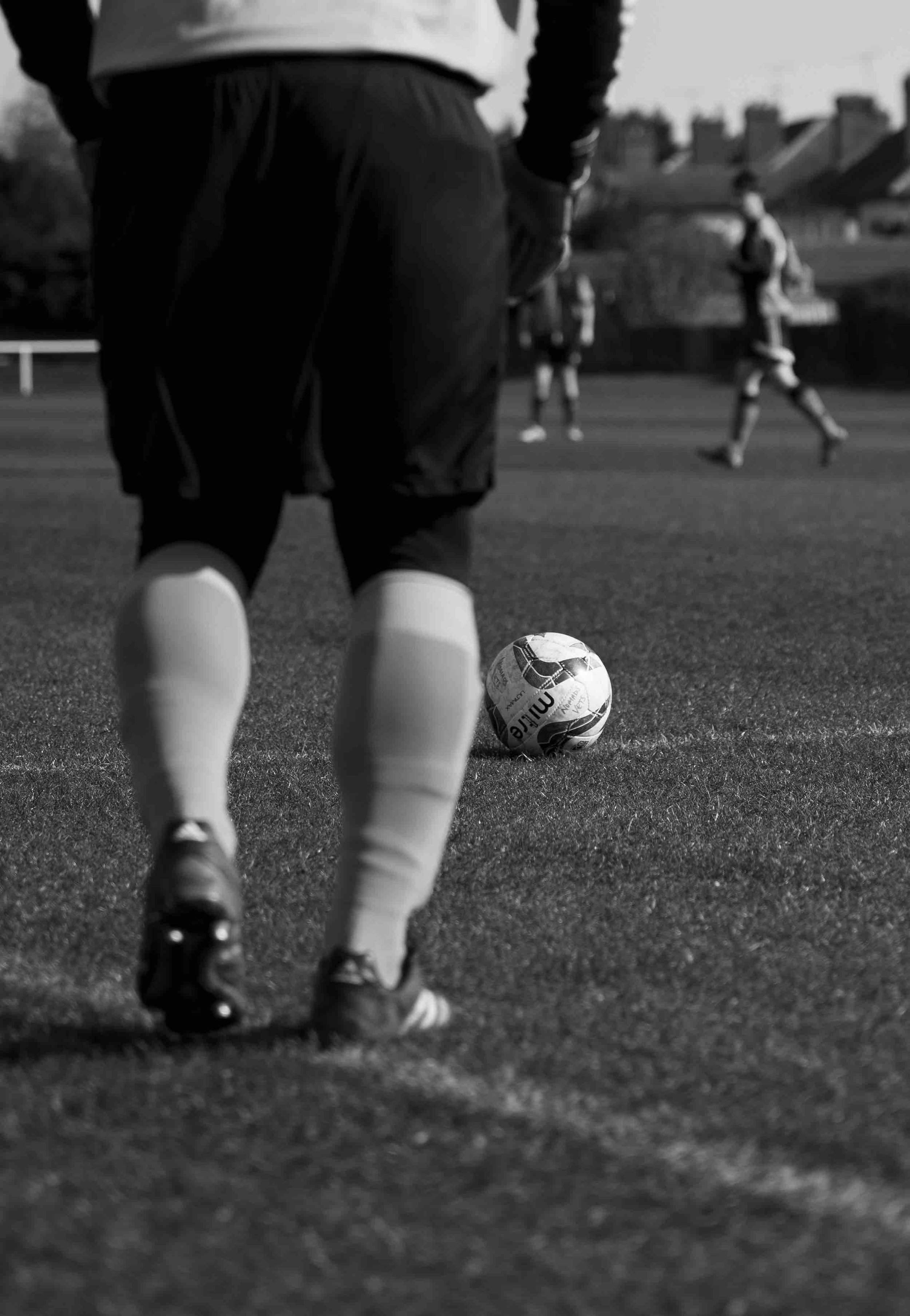 A soccer player standing near a ball on a field, with other players in the background.