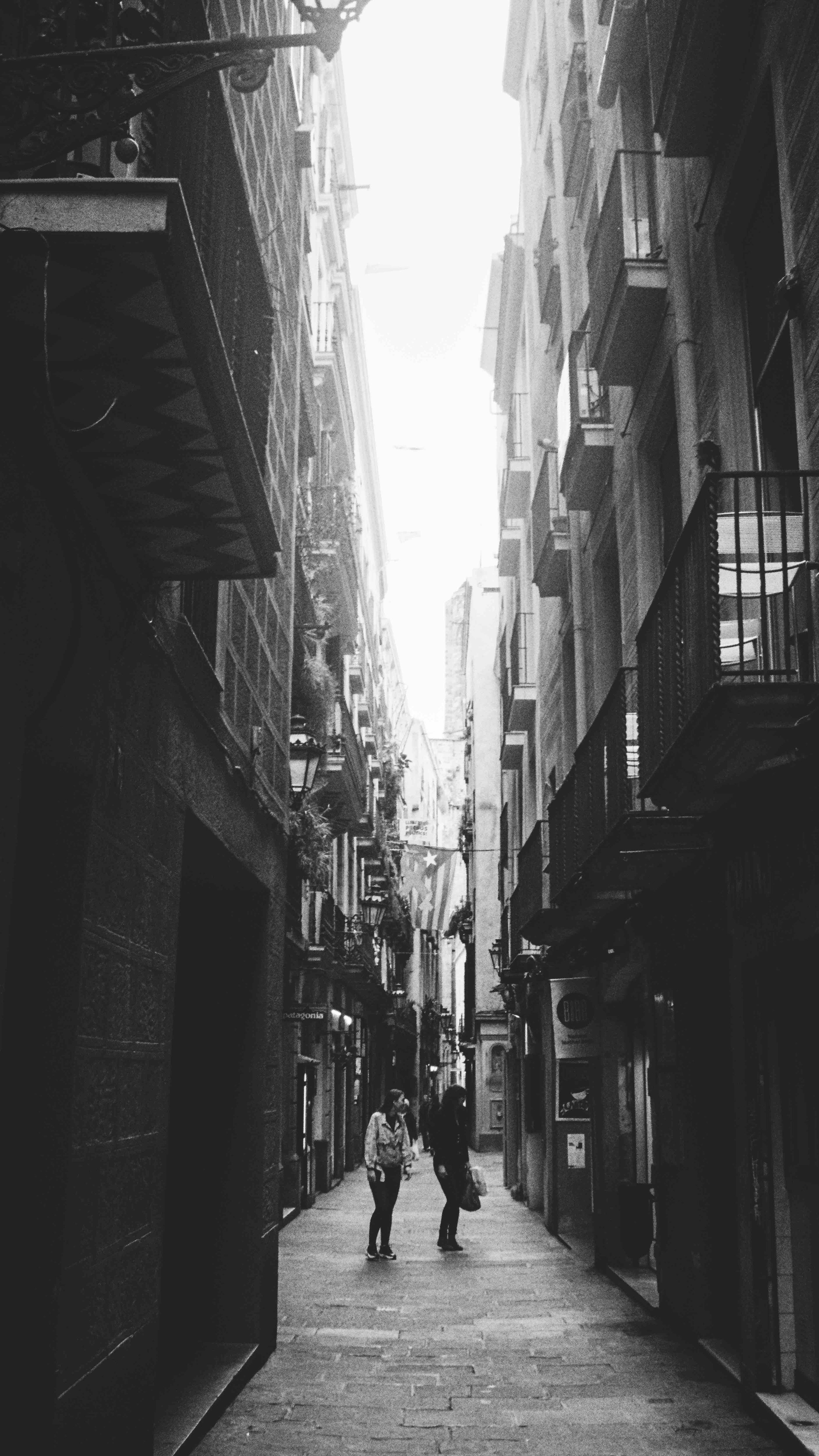 Black and white photo of a narrow European street, with two people walking, surrounded by tall buildings and balconies.