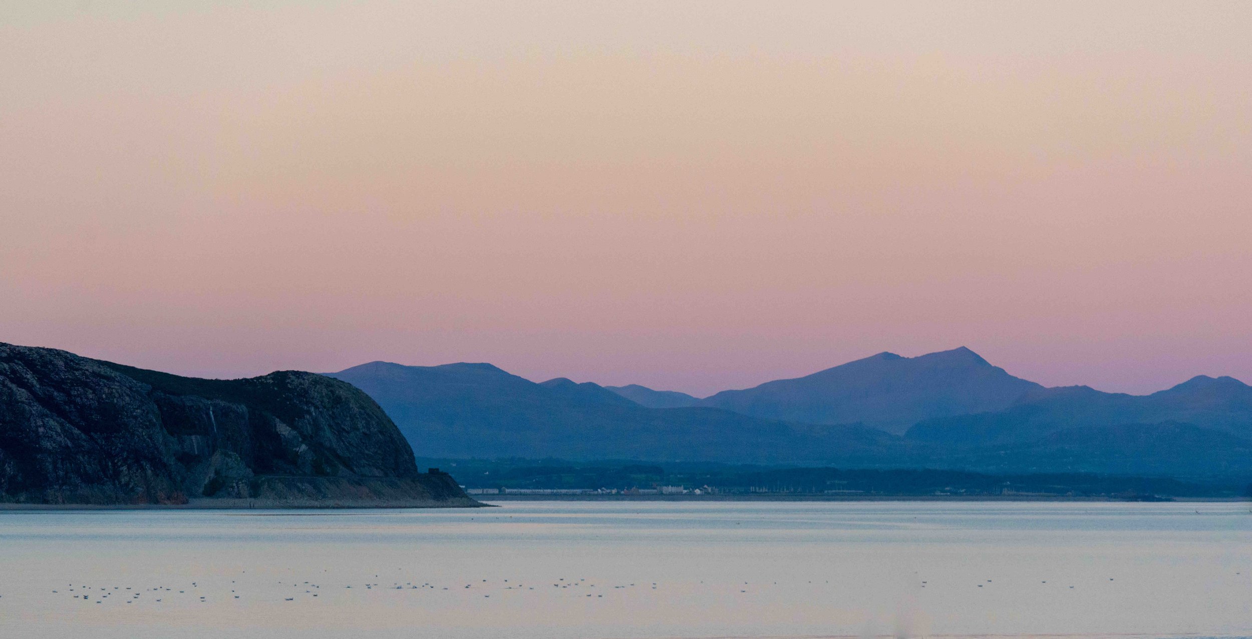 Coastal landscape with rocky cliffs and distant mountains at sunset, calm sea in foreground, sky with pink and blue hues.