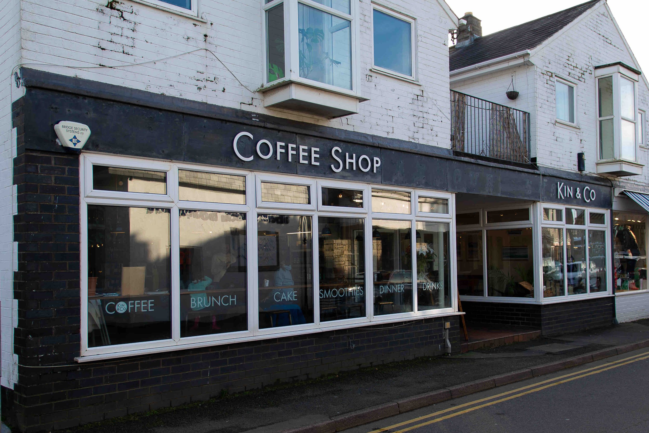 Exterior of a coffee shop named Kin & Co with large windows and signage for coffee, brunch, cake, smoothies, dinner, and drinks.