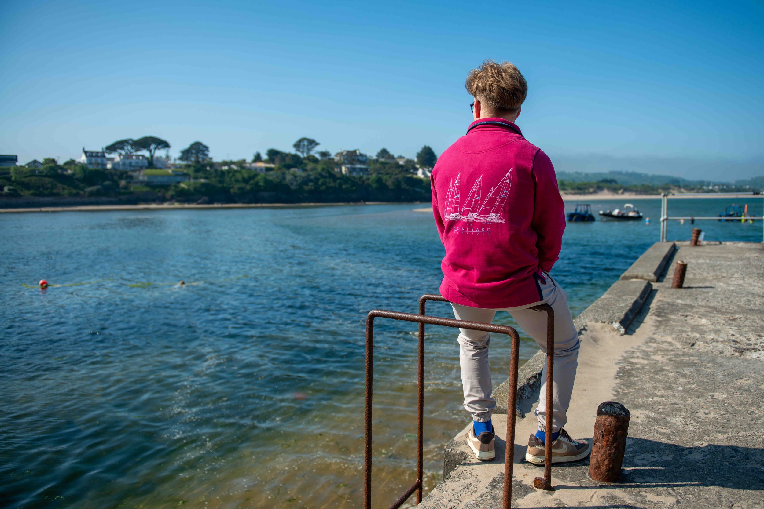 Person in a red jacket sitting on a pier overlooking a body of water with boats and distant shoreline under a clear blue sky.