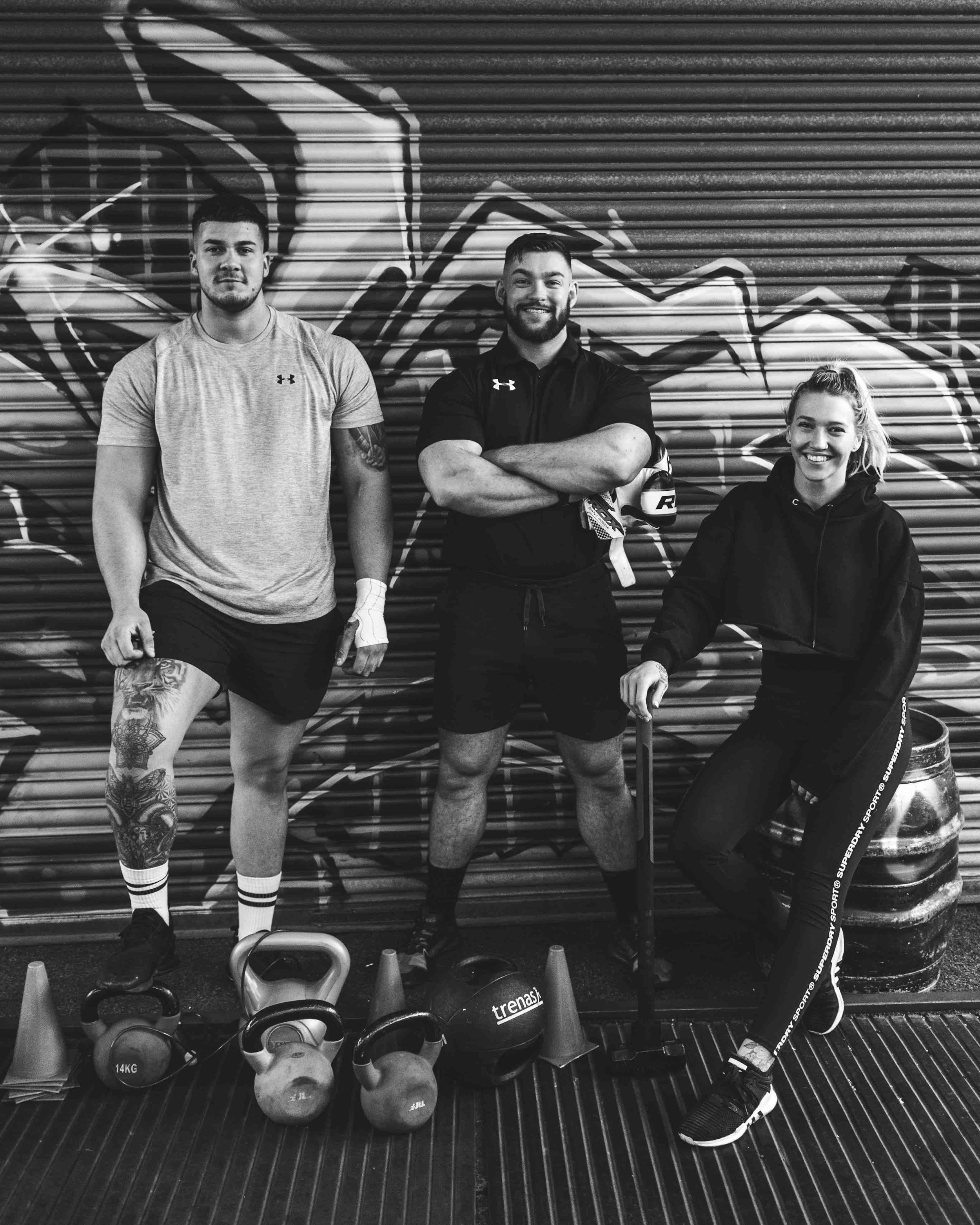 Three people posing with gym equipment in front of a graffiti-covered wall, including kettlebells and cones.