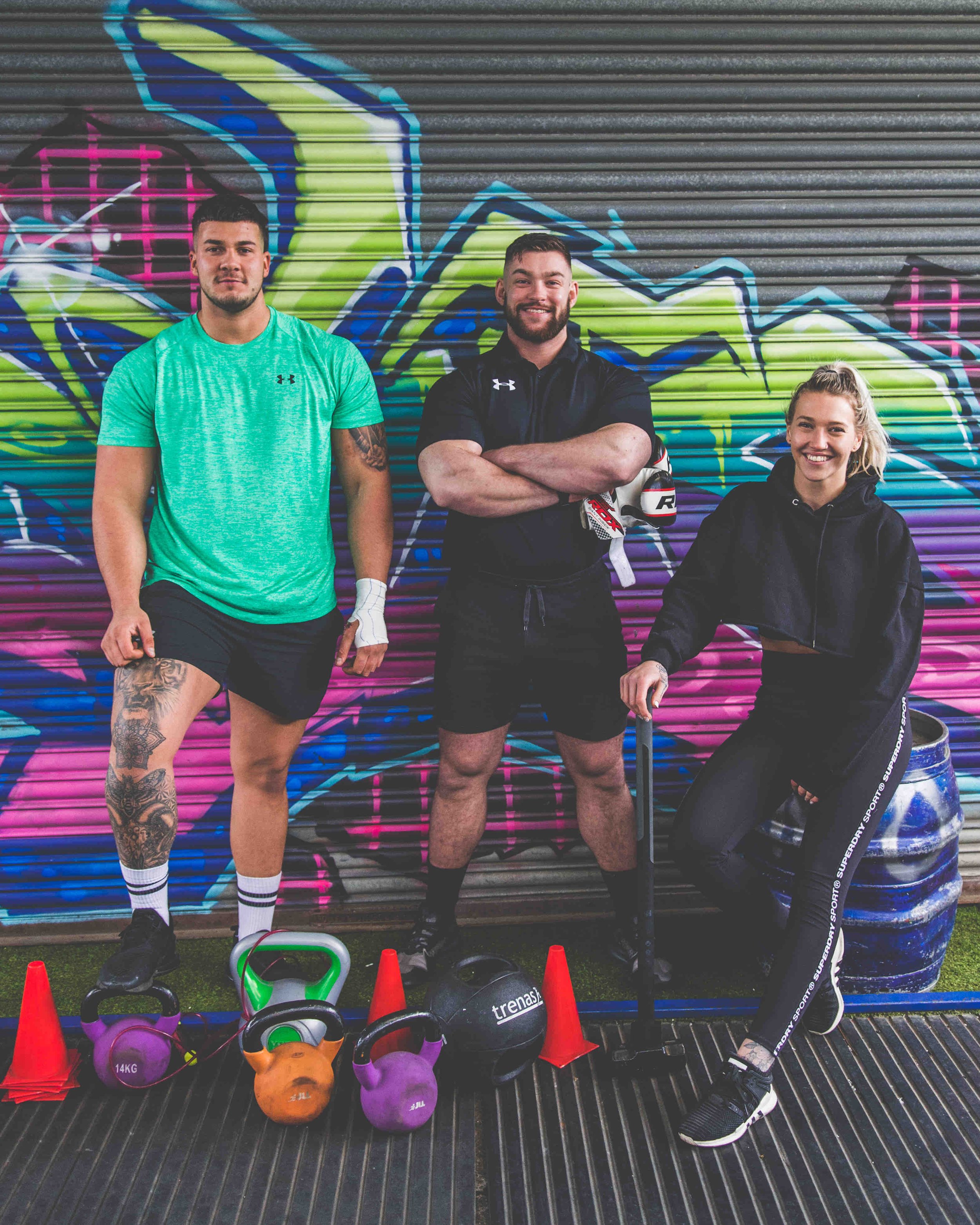 Three people standing in front of graffiti-covered background, with gym equipment like kettlebells and cones in front of them.
