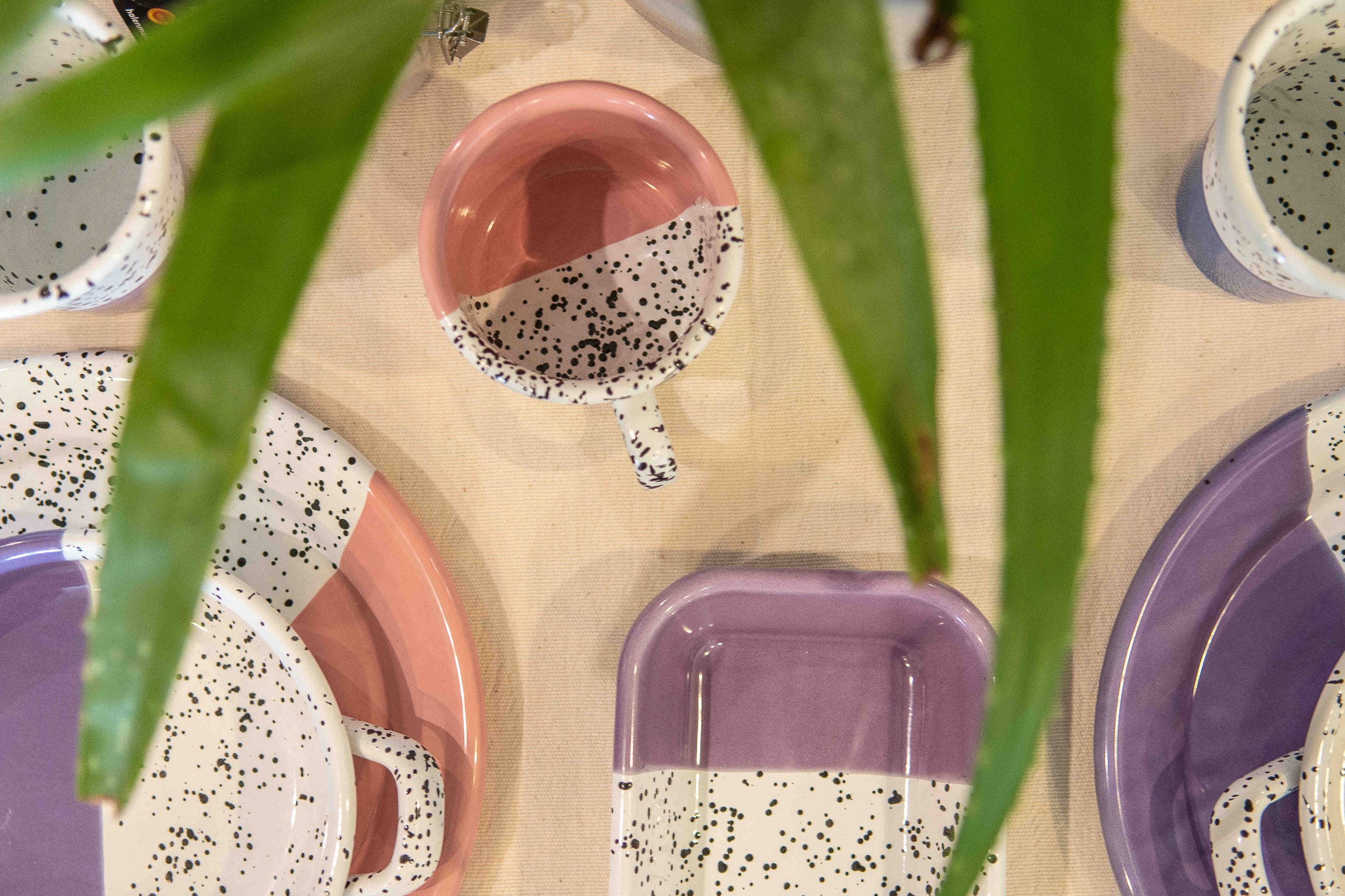 Overhead view of a collection of speckled ceramic tableware, including plates and cups in pastel colors, partially obscured by green leaves.