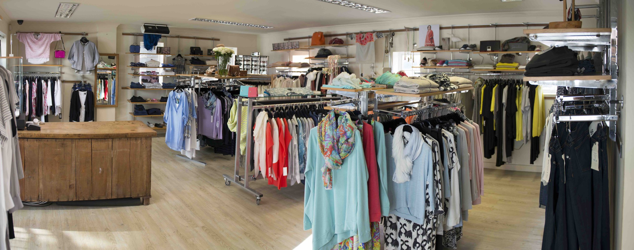 Interior of a boutique clothing store with racks of colorful clothes, a wooden checkout counter, and shelves displaying accessories.