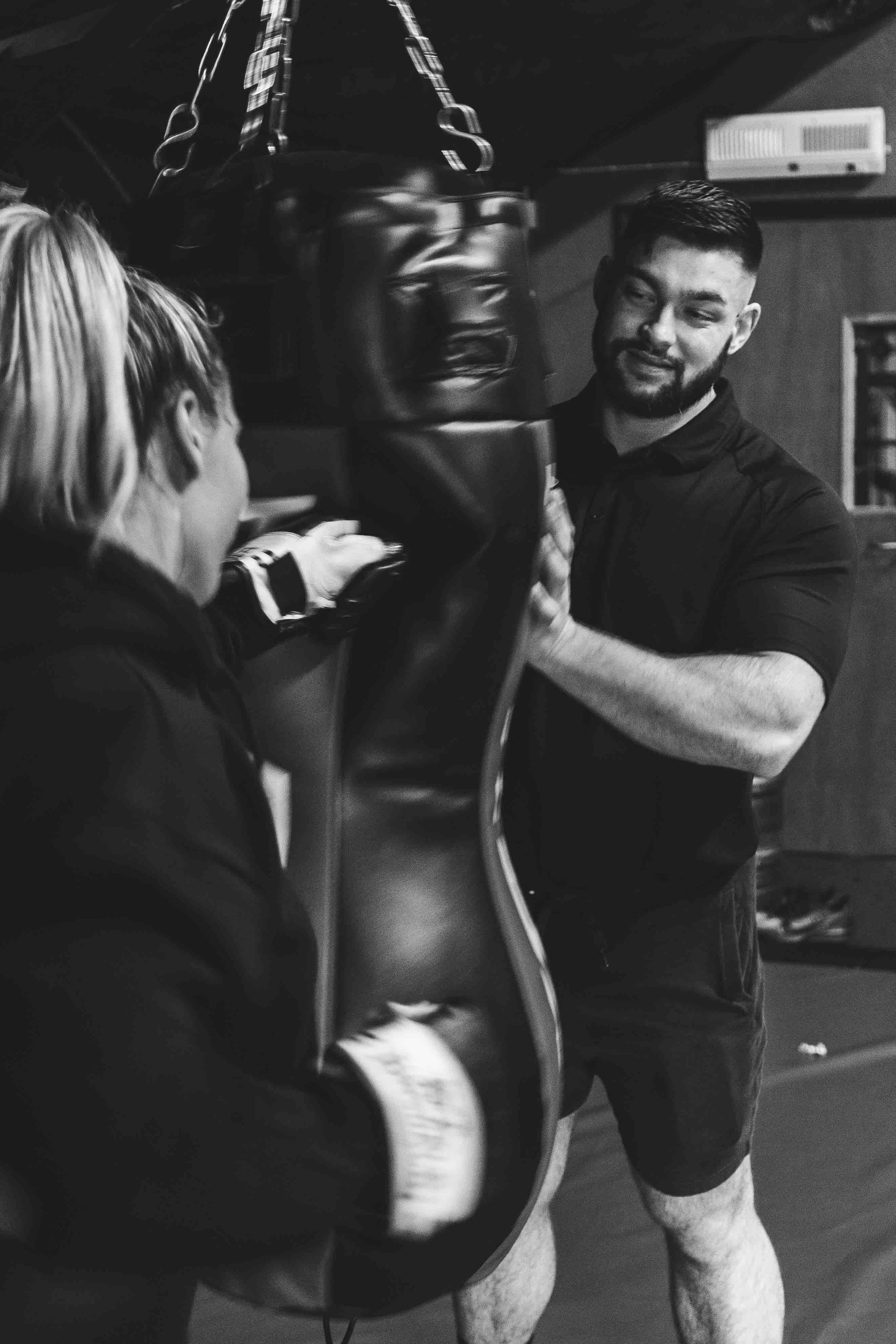 Black and white photo of a woman punching a boxing bag with a man holding it steady in a gym setting.