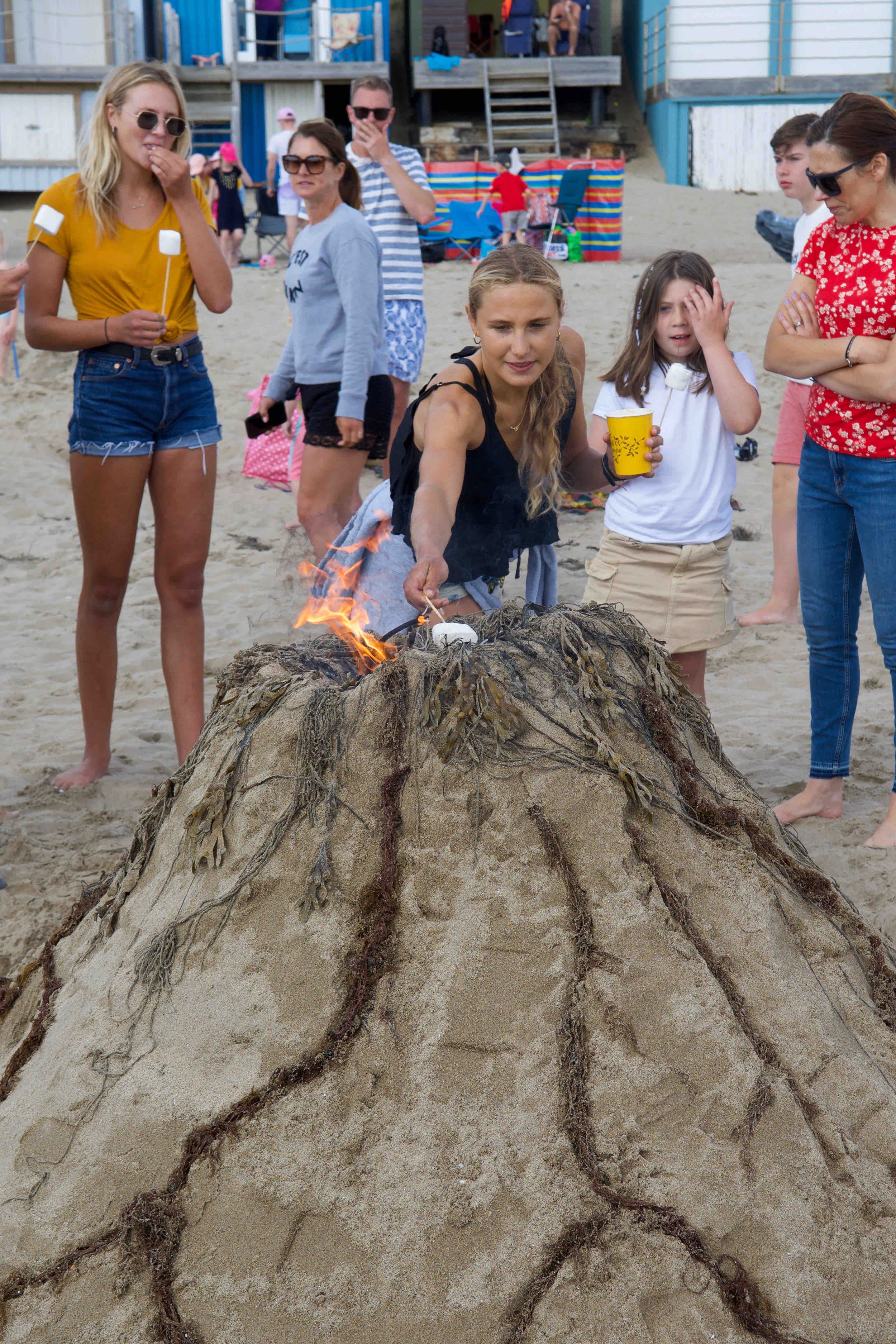 People gathered around a sand mound with fire on a beach, some holding sticks for toasting marshmallows.