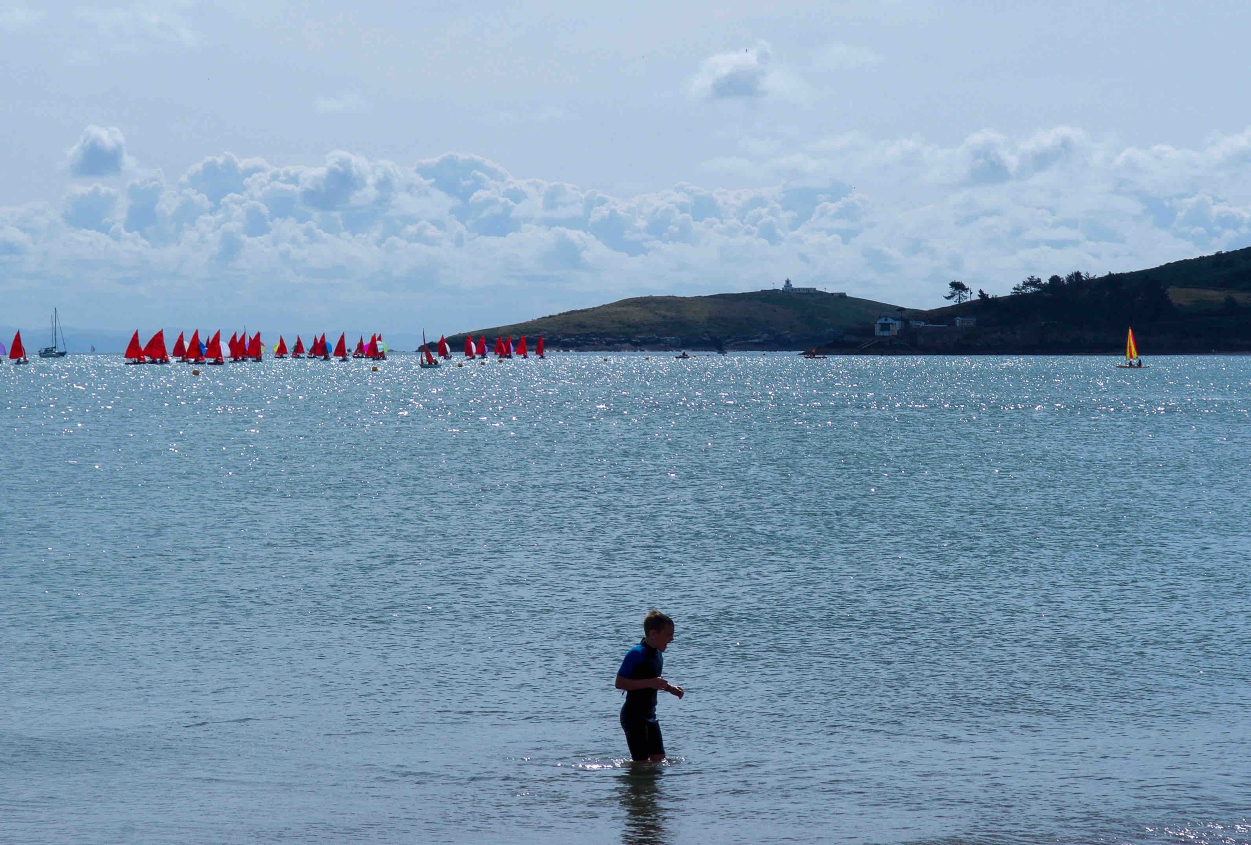 A person wading in shallow water near a beach, with many small sailboats with red sails on the distant water, and an island with light clouds in the background.