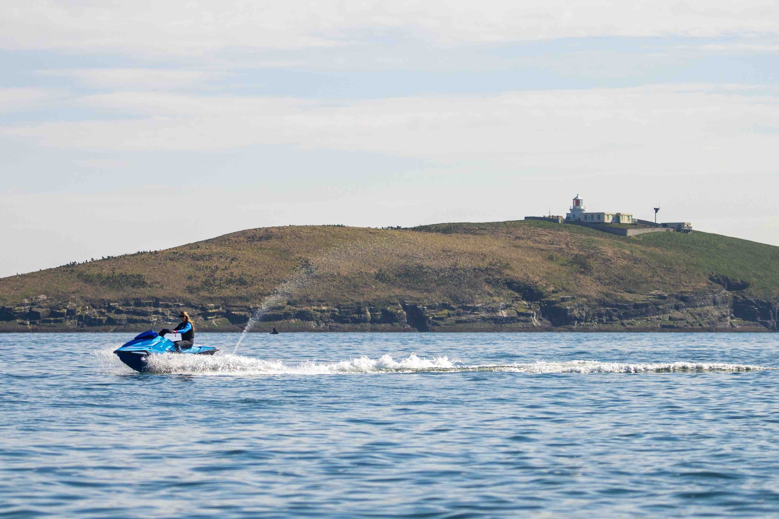 Person riding a blue jet ski on calm ocean water with a grassy island and lighthouse in the background.