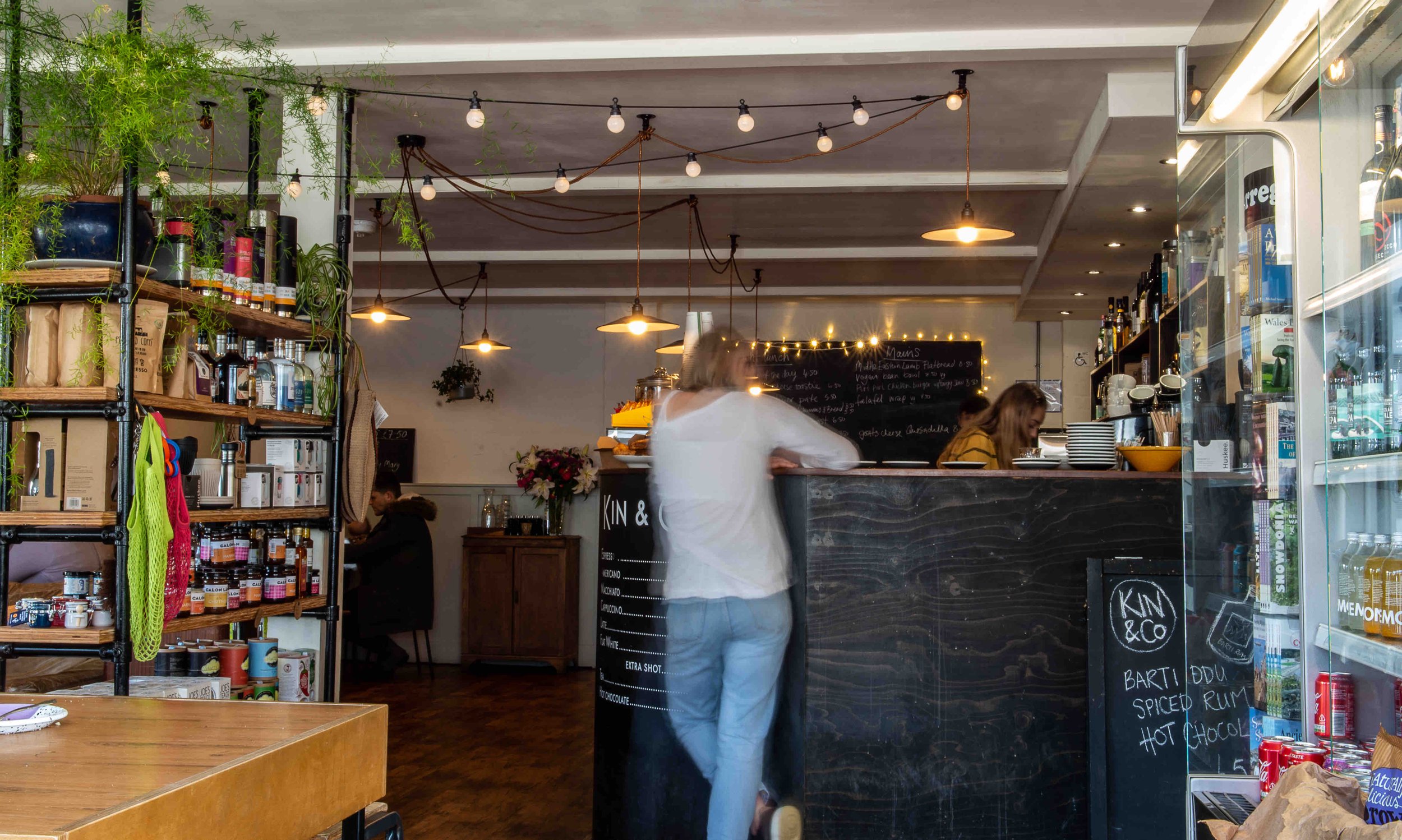 Cozy café interior with shelves of products, a person at the counter, hanging lights, and menu board.