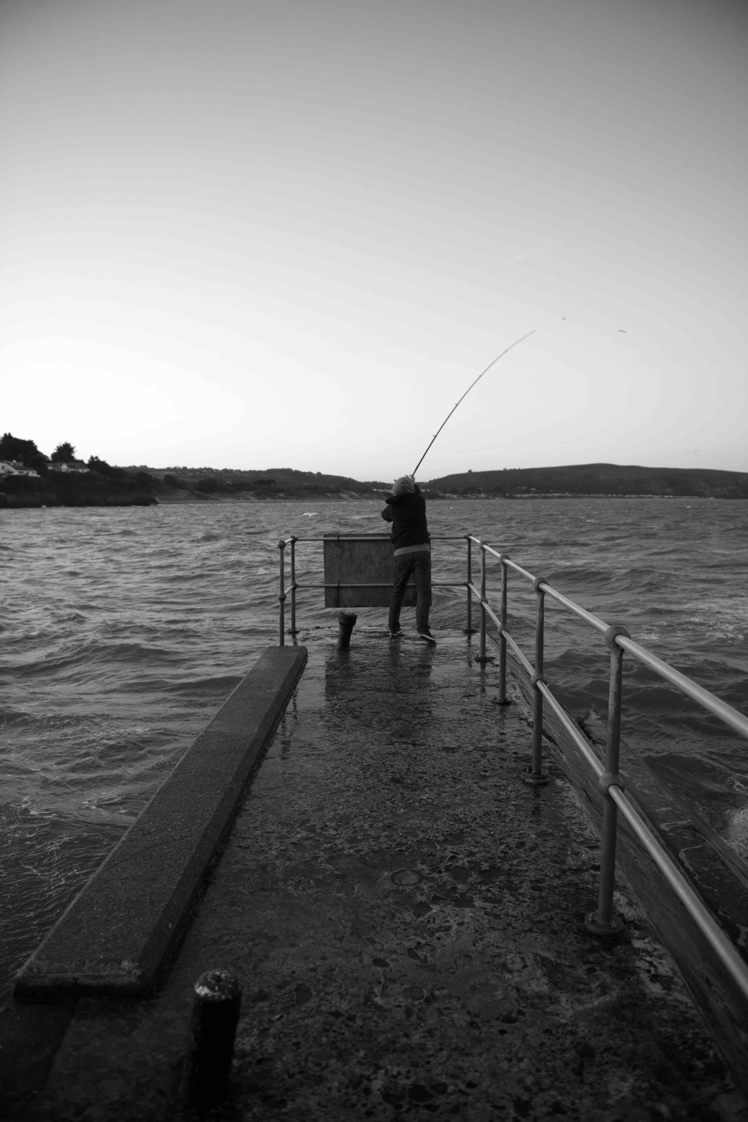 Black and white photo of a person fishing with a rod on a concrete pier overlooking a body of water, with hills and trees in the background.