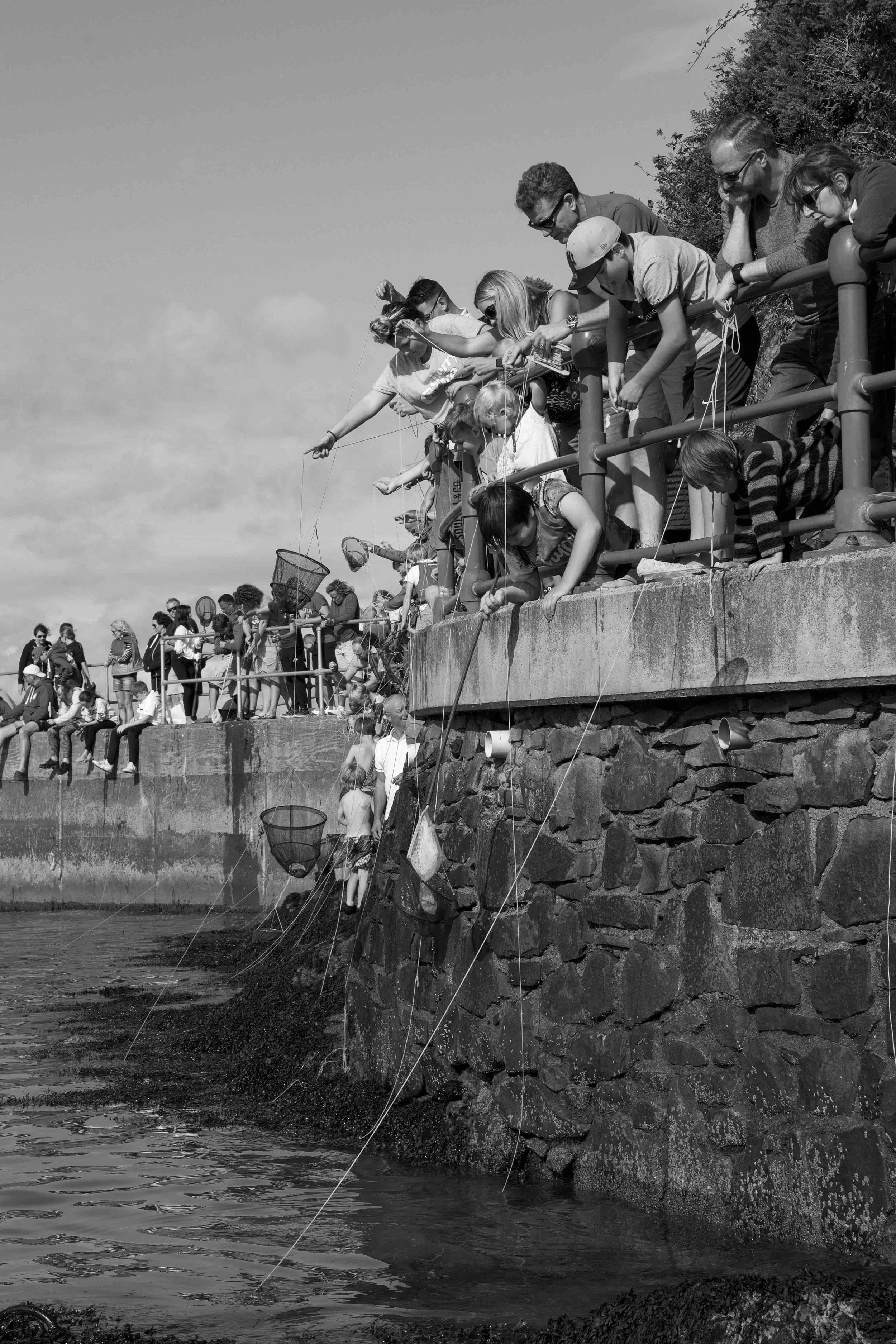People standing on a stone wall near the water, engaging in crabbing activities with nets and lines.
