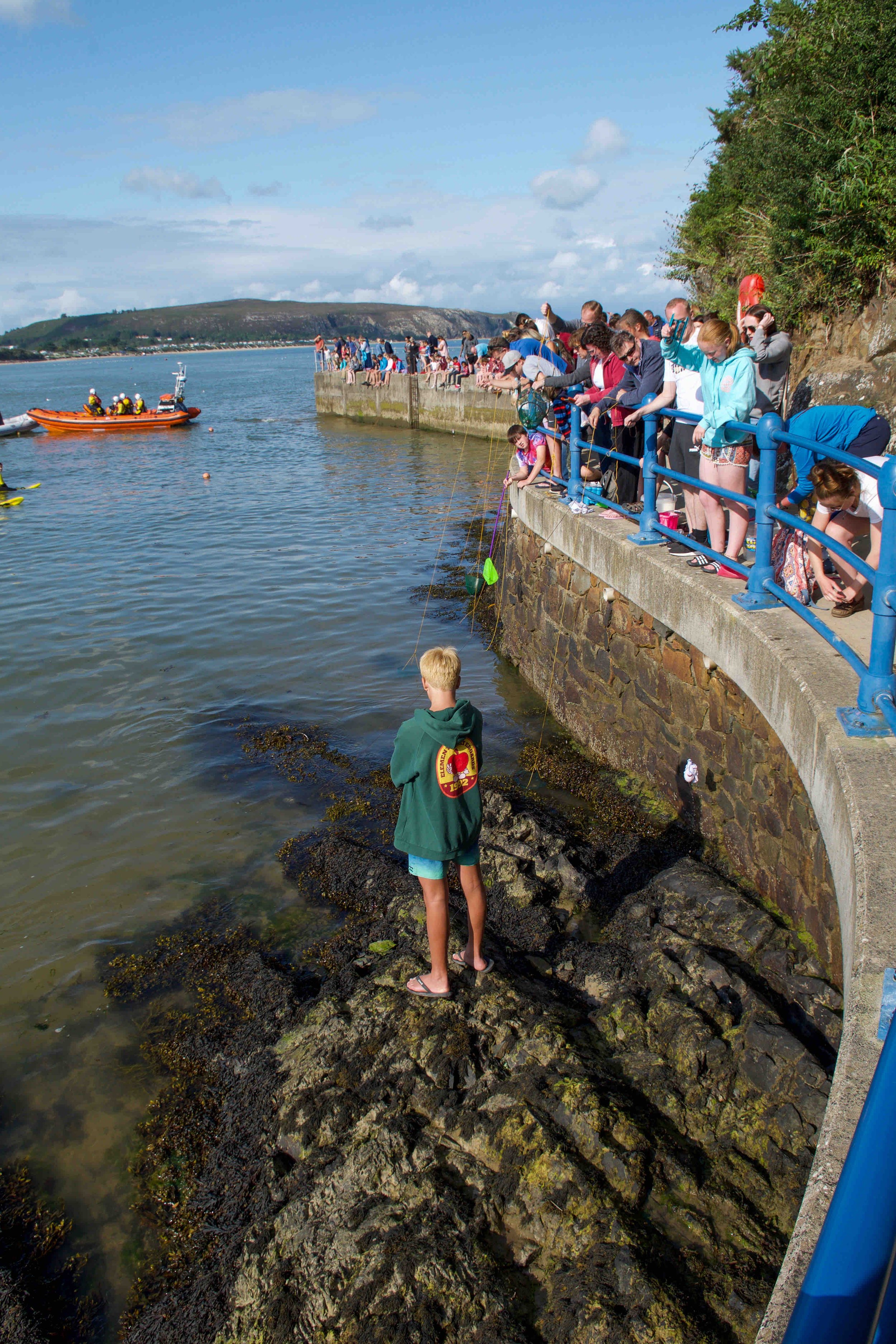 A group of people gathered on a sea wall, watching activities in the water below. Some people are fishing with lines dangling over the side. An orange rescue boat with crew members is nearby, and a person stands on rocky terrain near the shore. The s