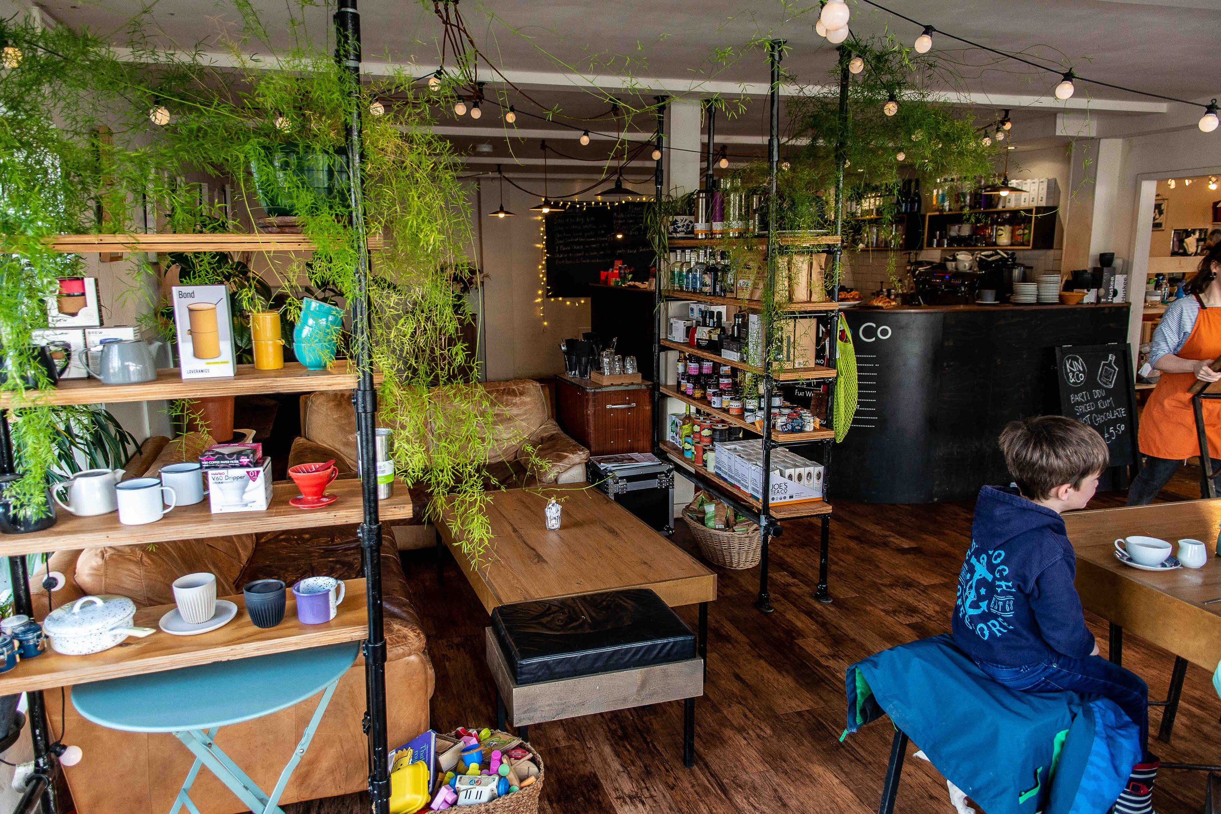 Interior of a cozy café with wooden shelves displaying mugs and kitchen items. There's a table with toys, a sofa, and a child sitting at a table. Green plants hang from the ceiling, adding a natural touch. The counter area with drinks and products is