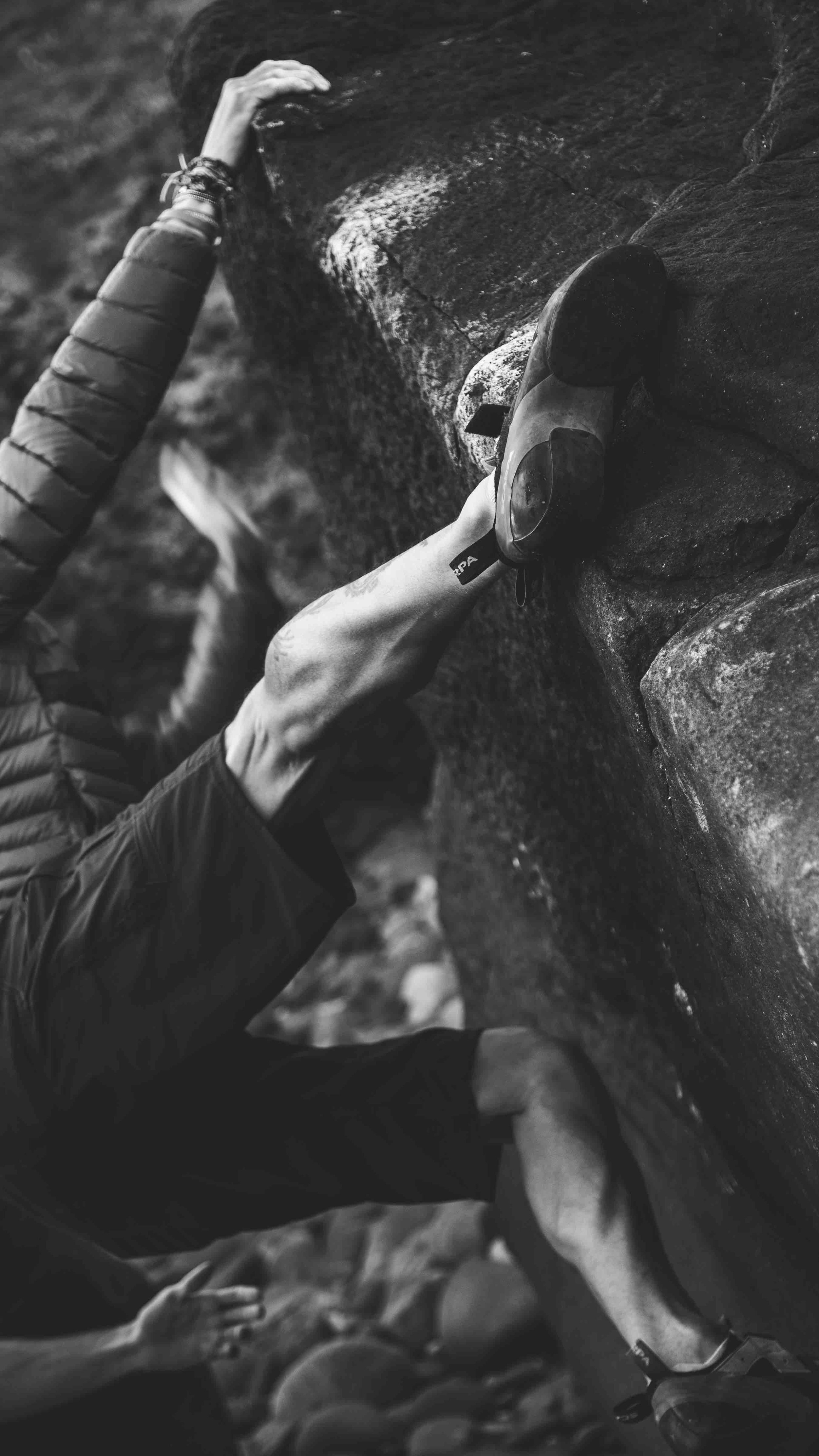 Black and white photo of a rock climber scaling a cliff, wearing climbing shoes and a jacket, with a focus on their hands and legs.