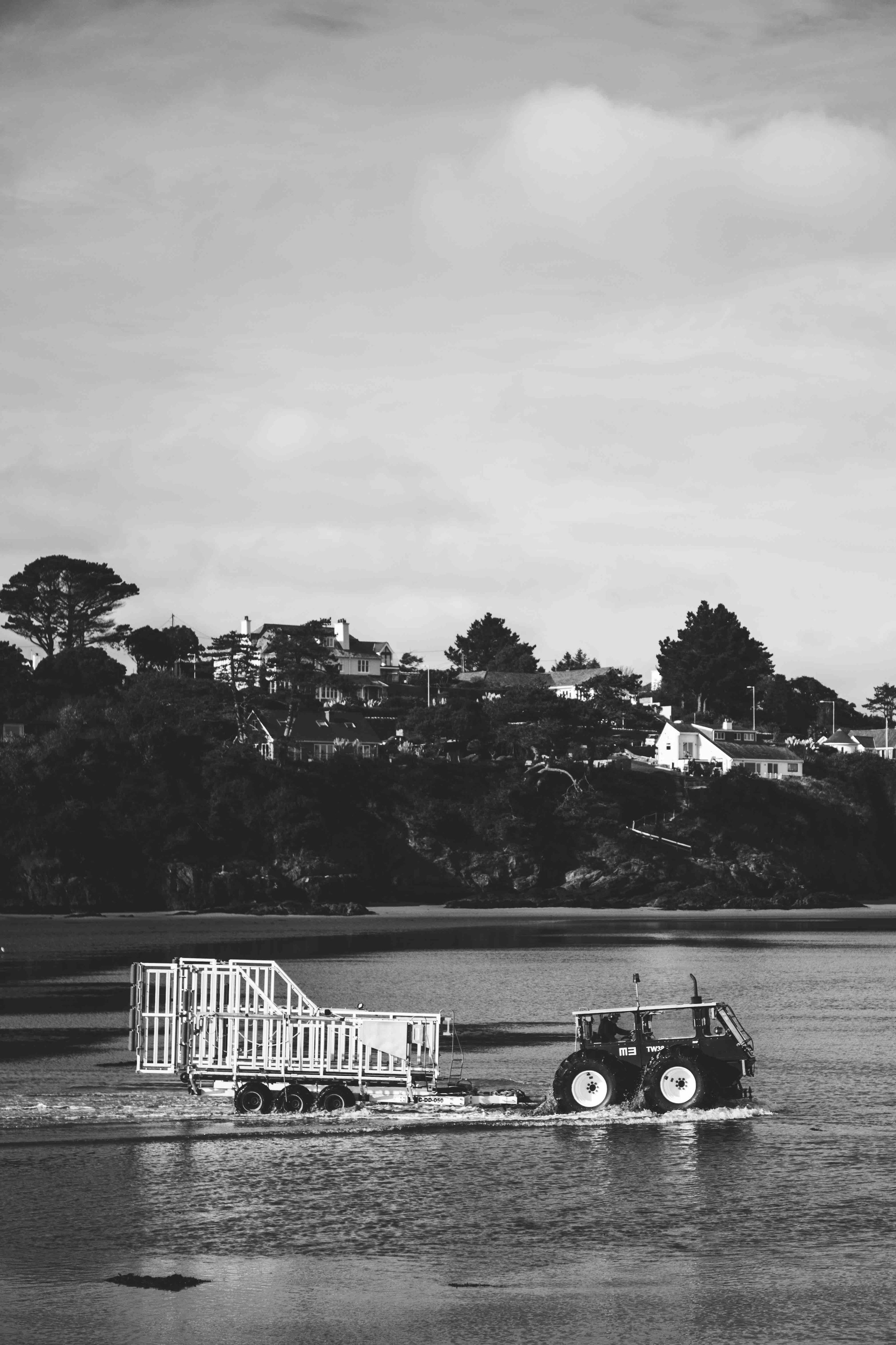 A black and white image of a tractor pulling a trailer through shallow water, with houses and trees on a hillside in the background.