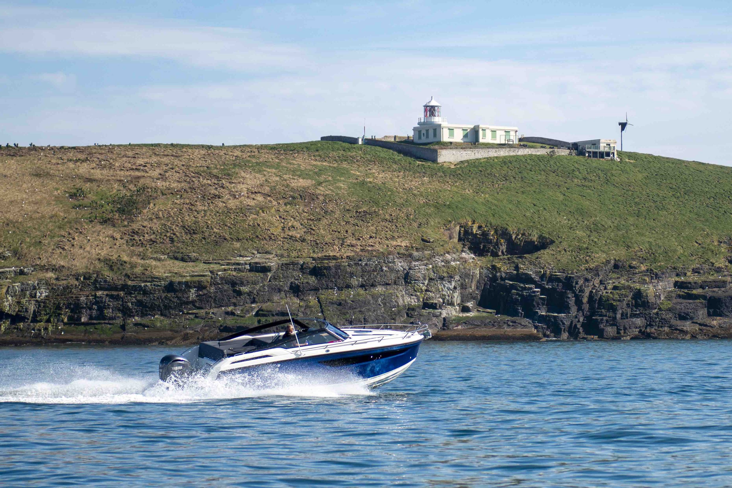 A speedboat moving on water with a grassy cliff and a lighthouse in the background on a clear day.