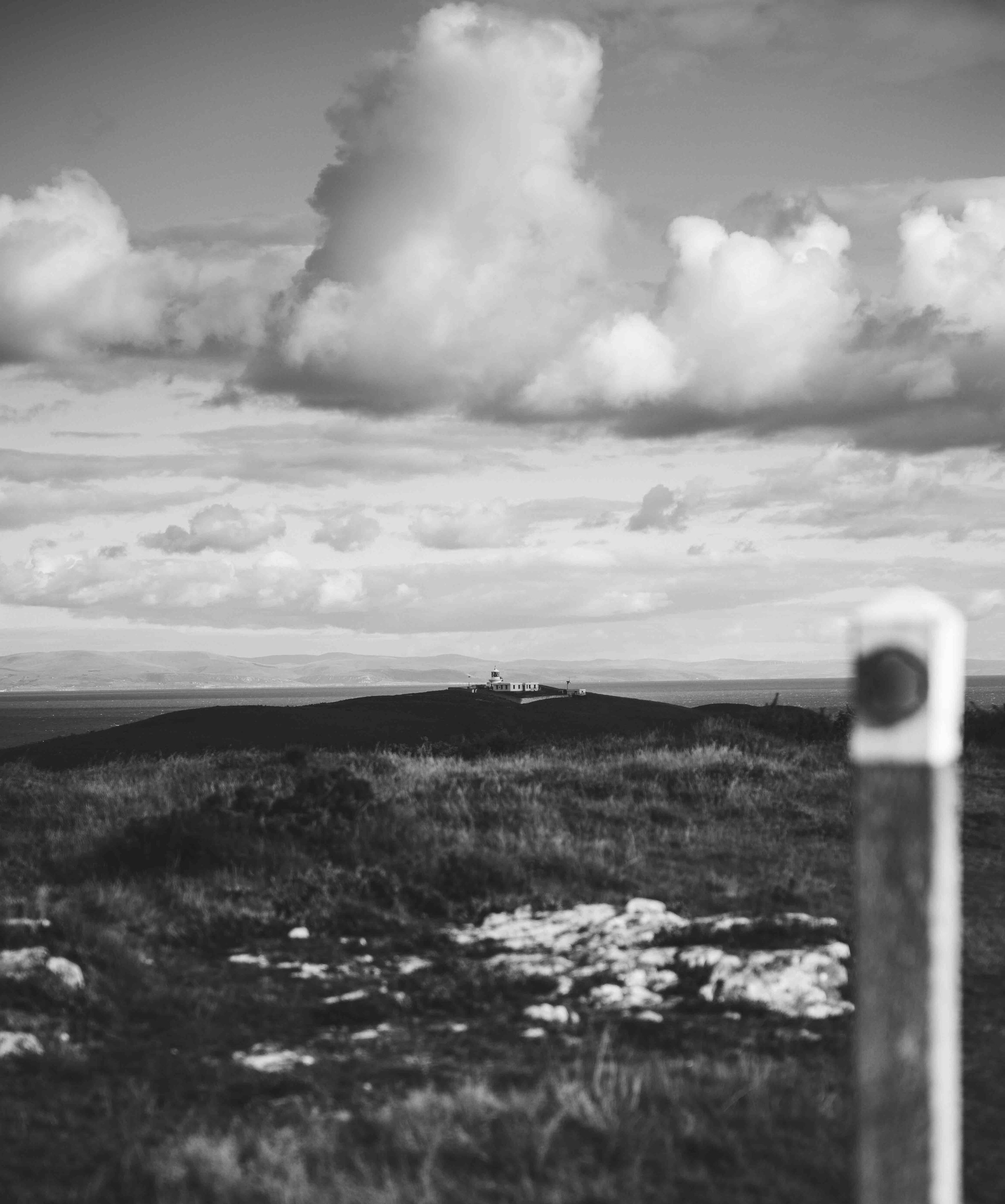 Black and white photo of a distant house on a hill, under a cloudy sky, with grass and rocks in the foreground.