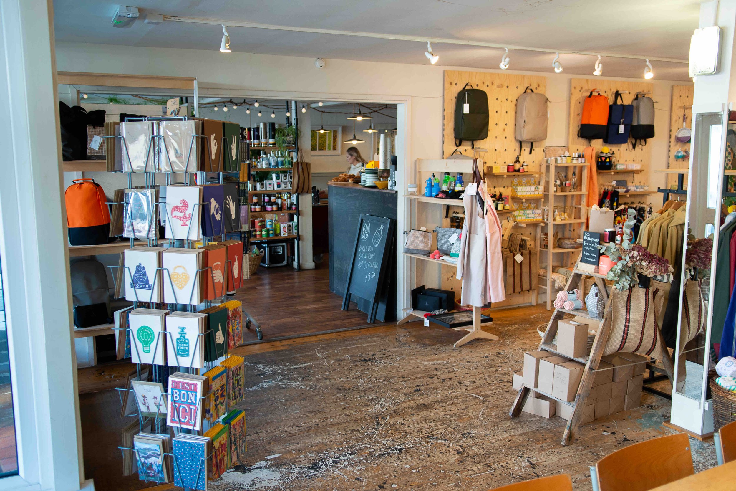 Interior of a small boutique store with greeting cards displayed on a rotating stand, backpacks on the wall, shelves with various products, and decorative items like boxes and dried flowers. A chalkboard sign stands near a counter in the background.