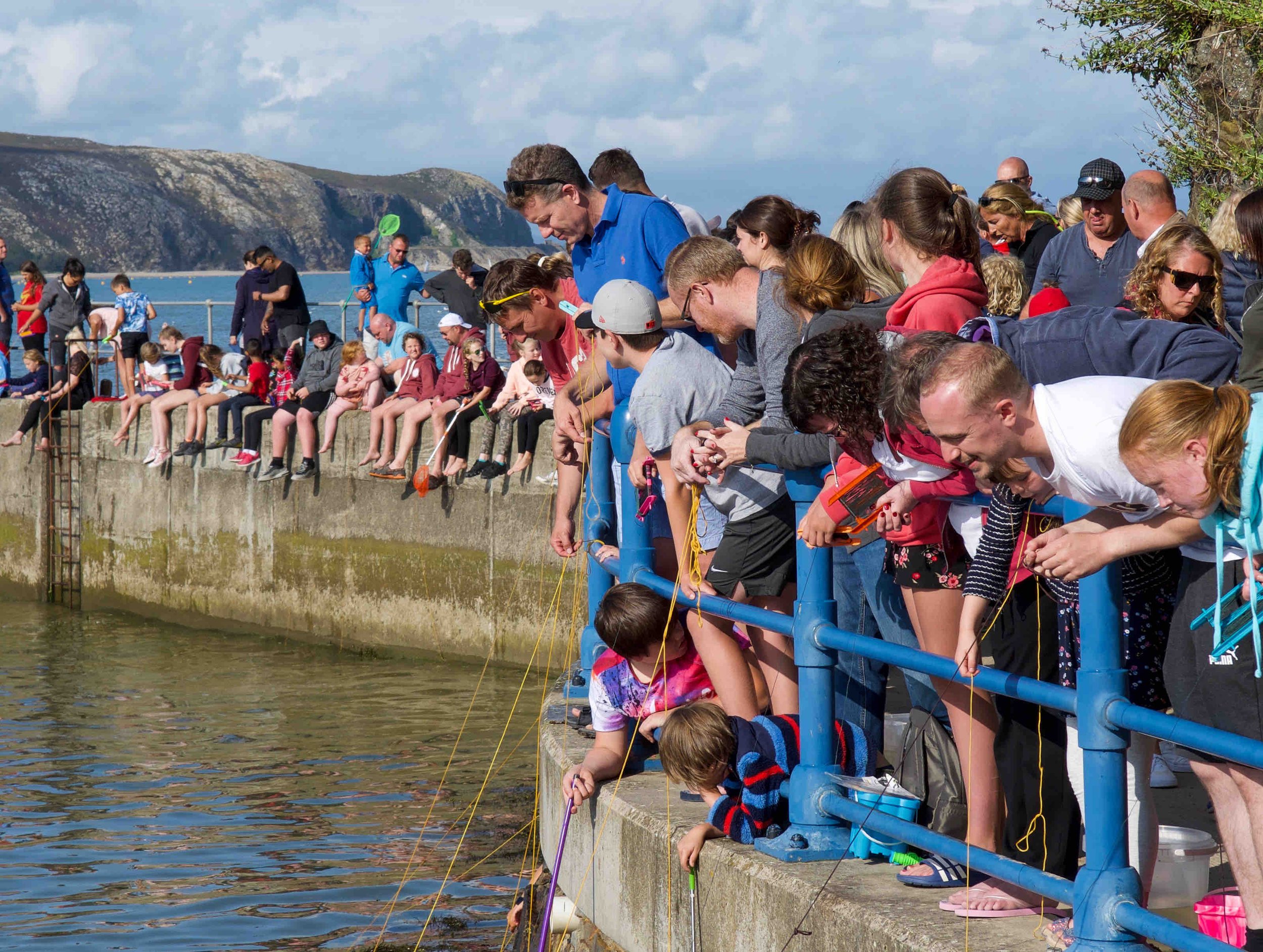 A group of people, including children and adults, gathered on a concrete pier overlooking water, engaged in activities like crabbing or fishing. A scenic coastline with hills is visible in the background under a blue sky with clouds.