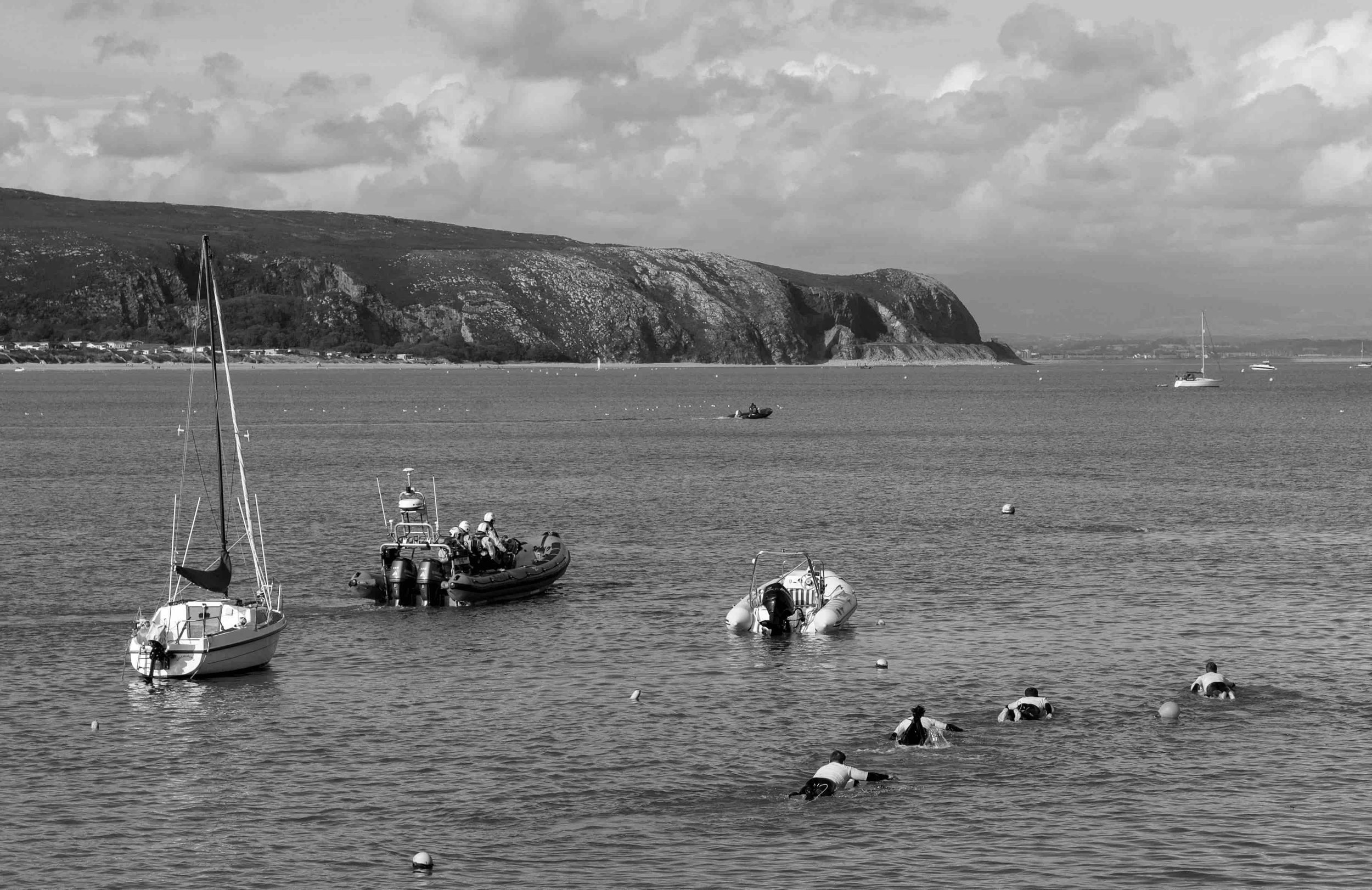 Boats and people swimming in a bay with cliffs in the background.