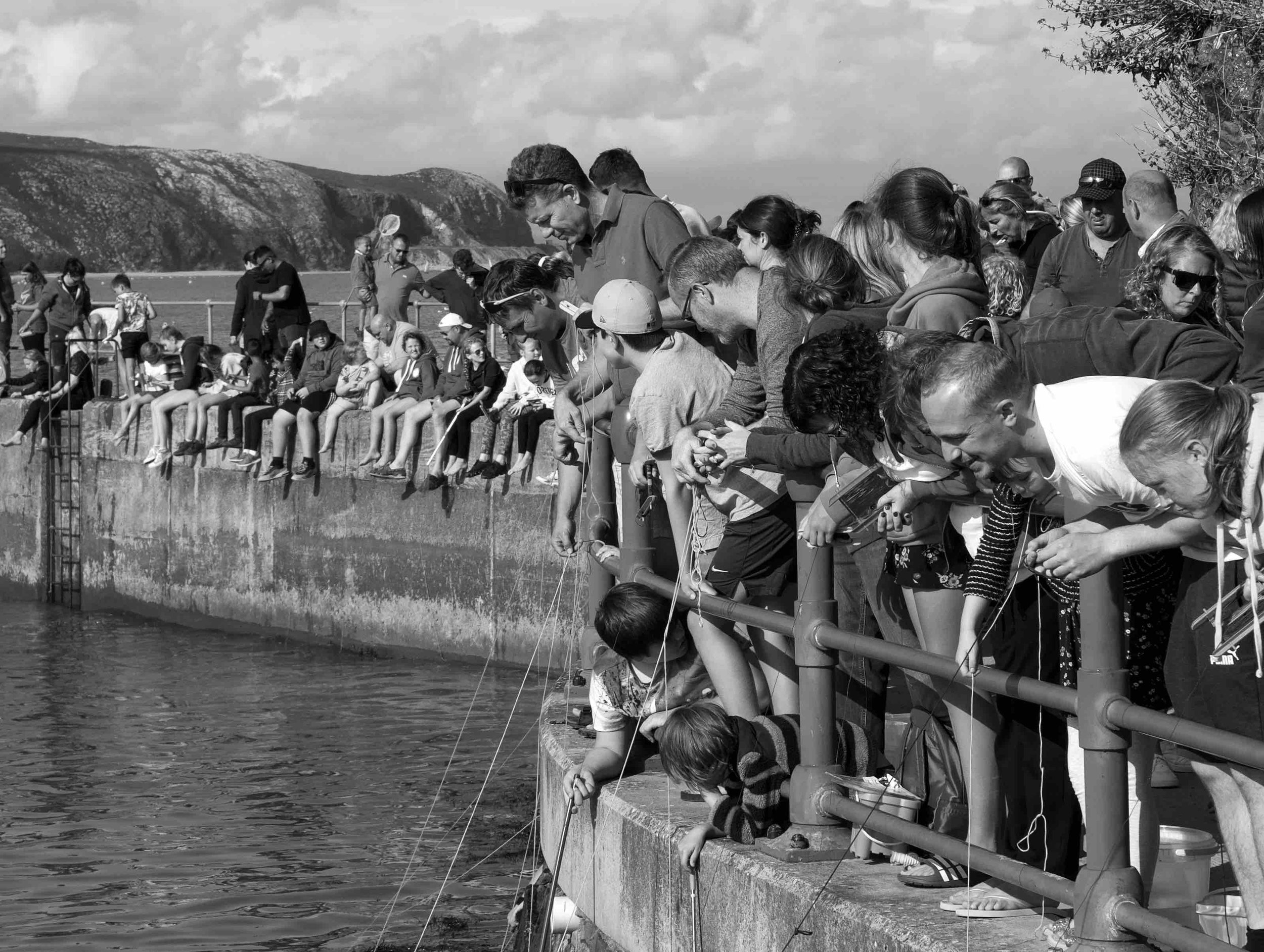 Black and white photo of people crab fishing off a harbor wall. A crowd of people, including children and adults, lean over the edge while holding strings into the water. Others sit on the wall observing.