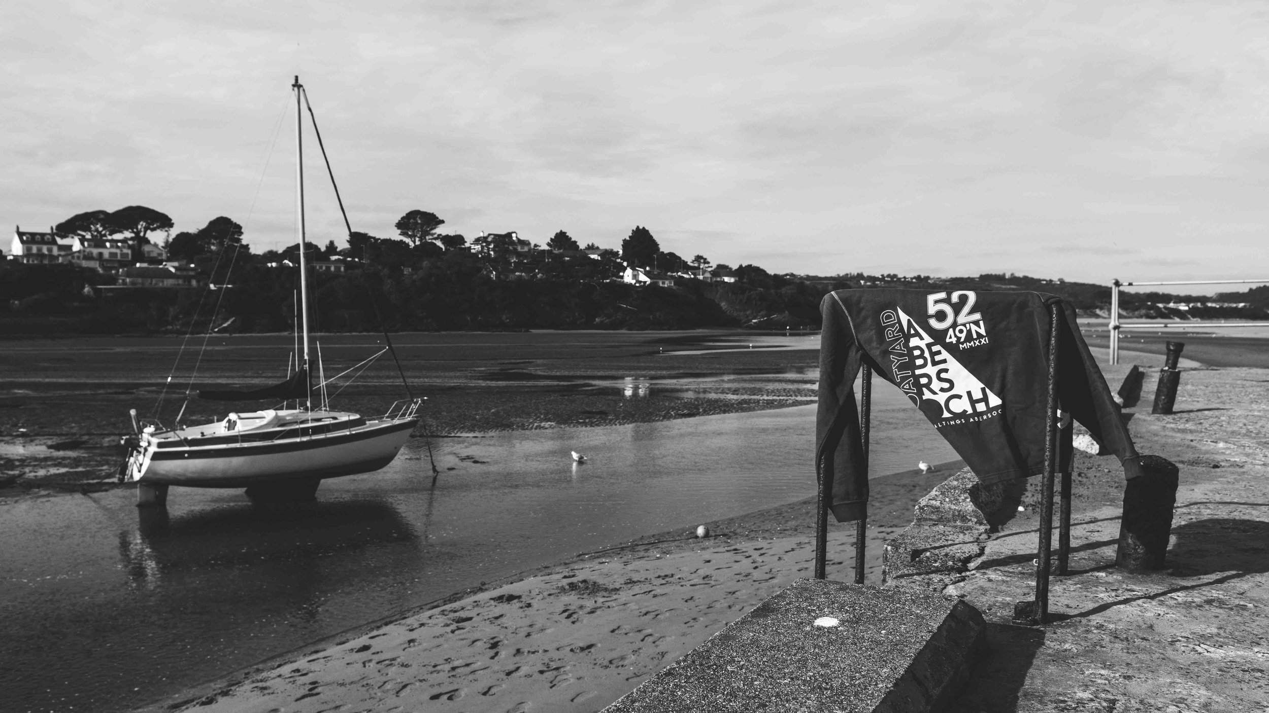 Black and white photo of a sailboat resting on a sandy shoreline during low tide, with a sweater hanging on a handrail in the foreground; houses and trees visible in the background.