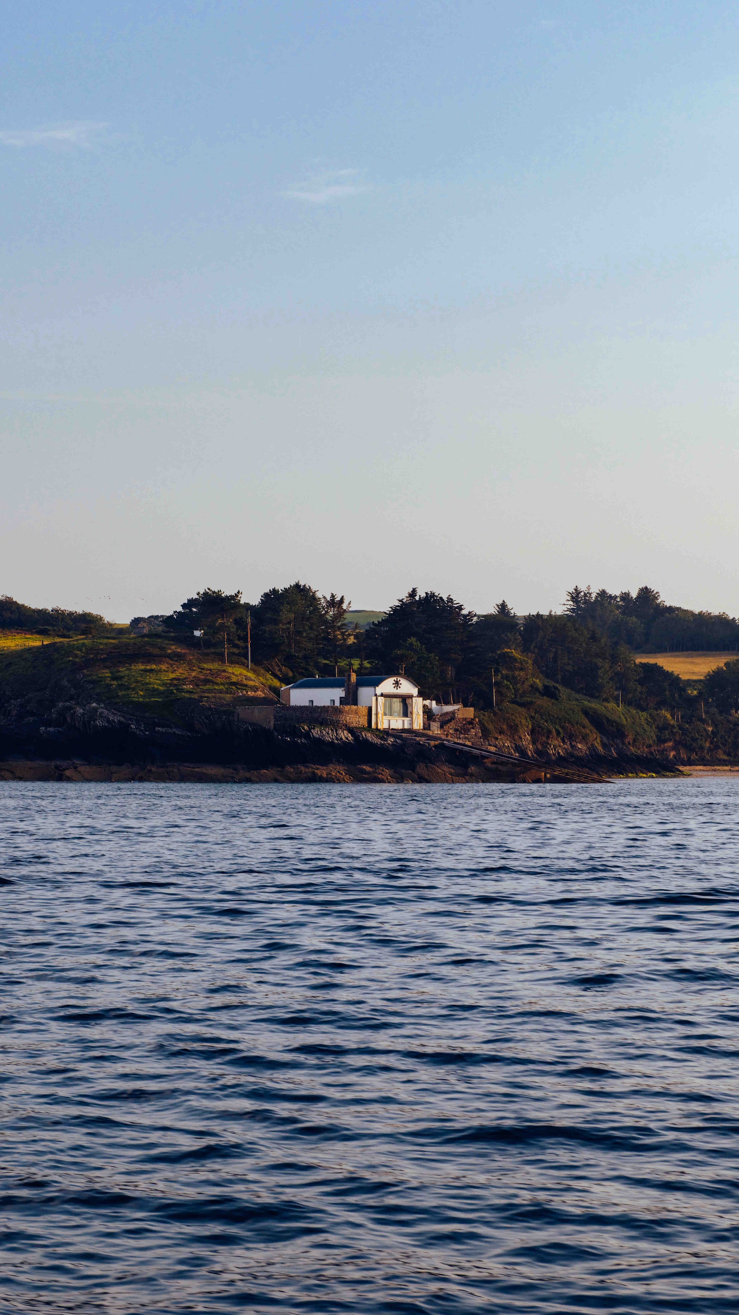 Coastal landscape with house on rocky hill, surrounded by trees and ocean.