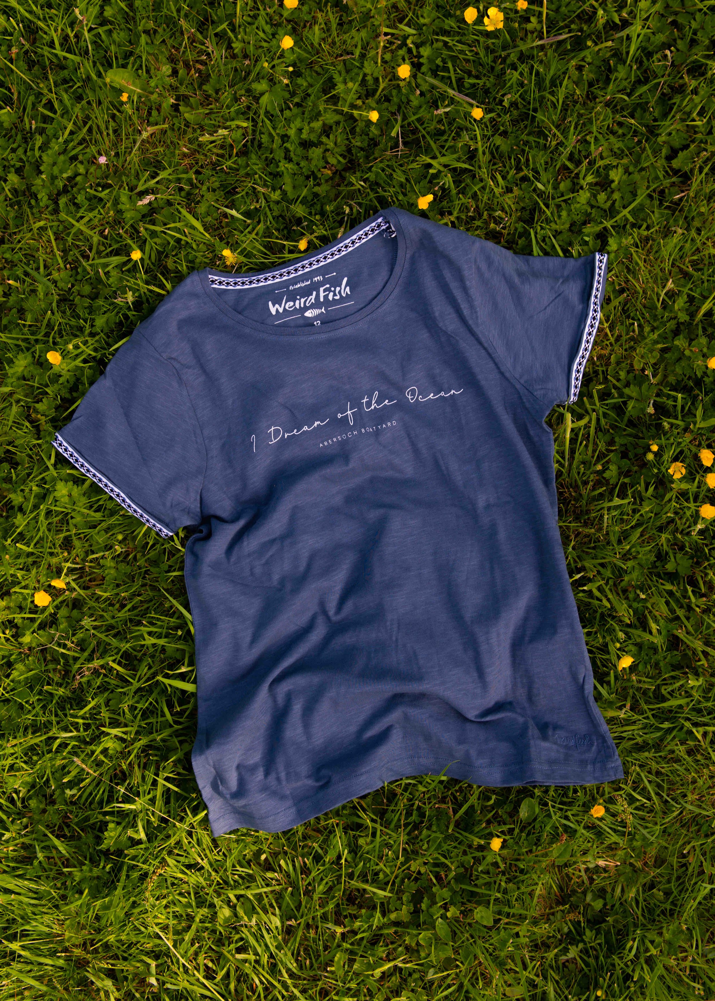Blue t-shirt with 'Weird Fish' label and ocean-themed text lying on green grass with yellow flowers.