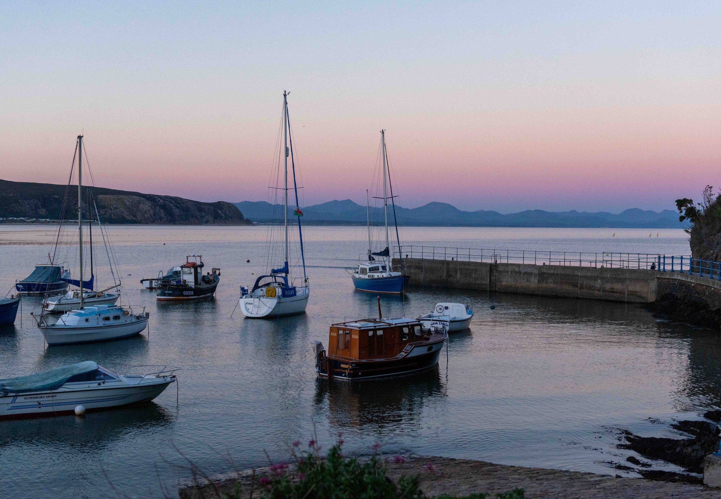 Small harbor with various boats at sunset, calm water, and distant mountains under a colorful sky.