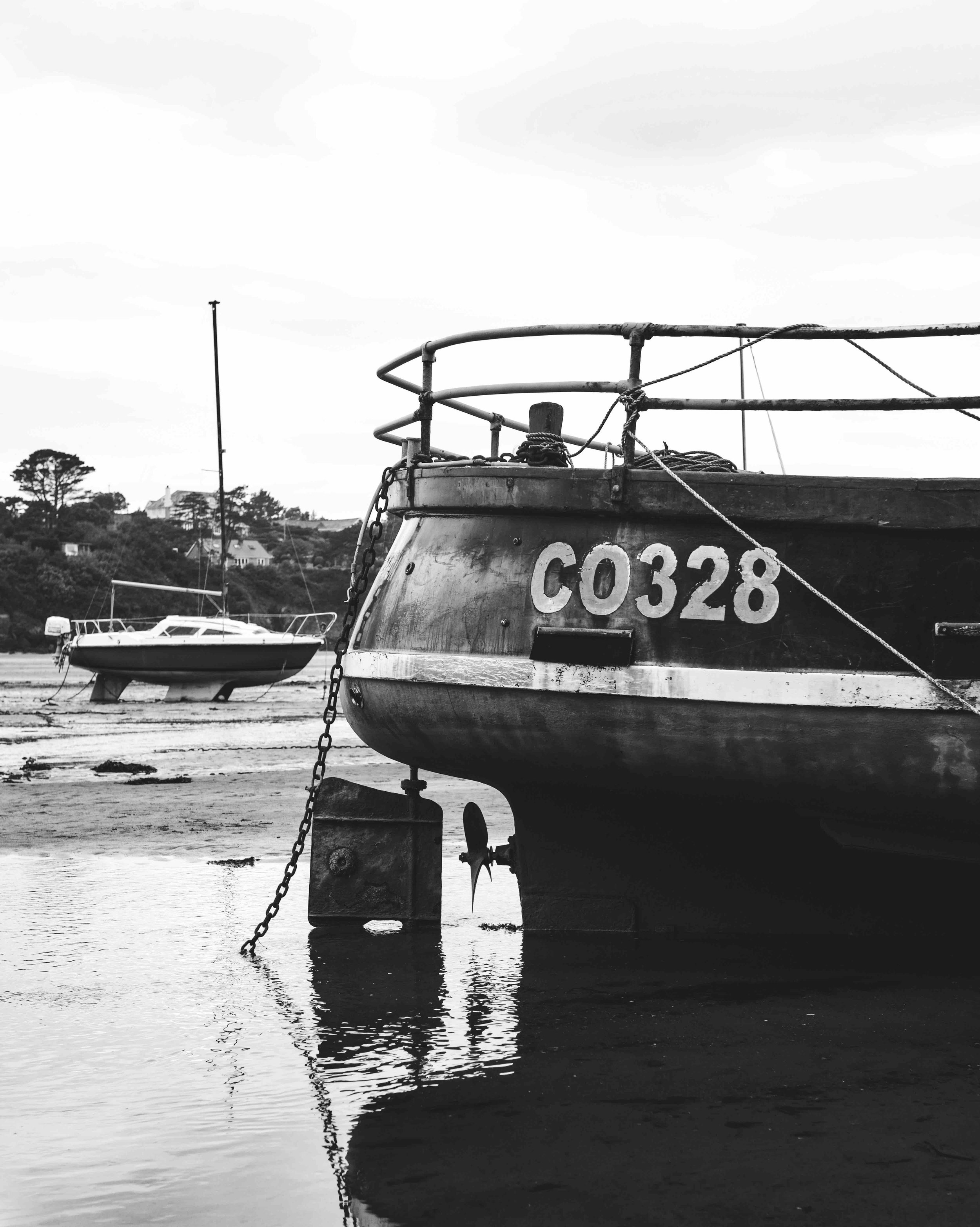 Black and white photo of a boat with the number C0328. The boat is moored on shallow water with another boat in the background, near trees and a building on the shore.