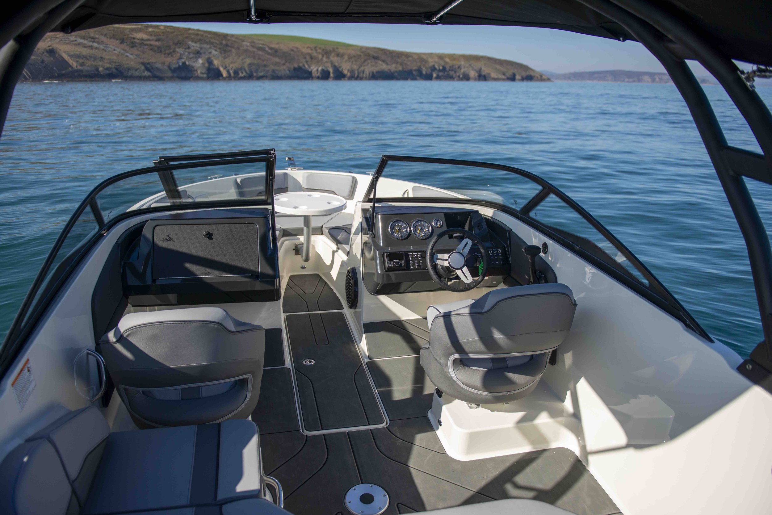 Interior view of a boat on the water with a steering wheel, seating area, and control panel under a canopy. Rocky shoreline is visible in the background.