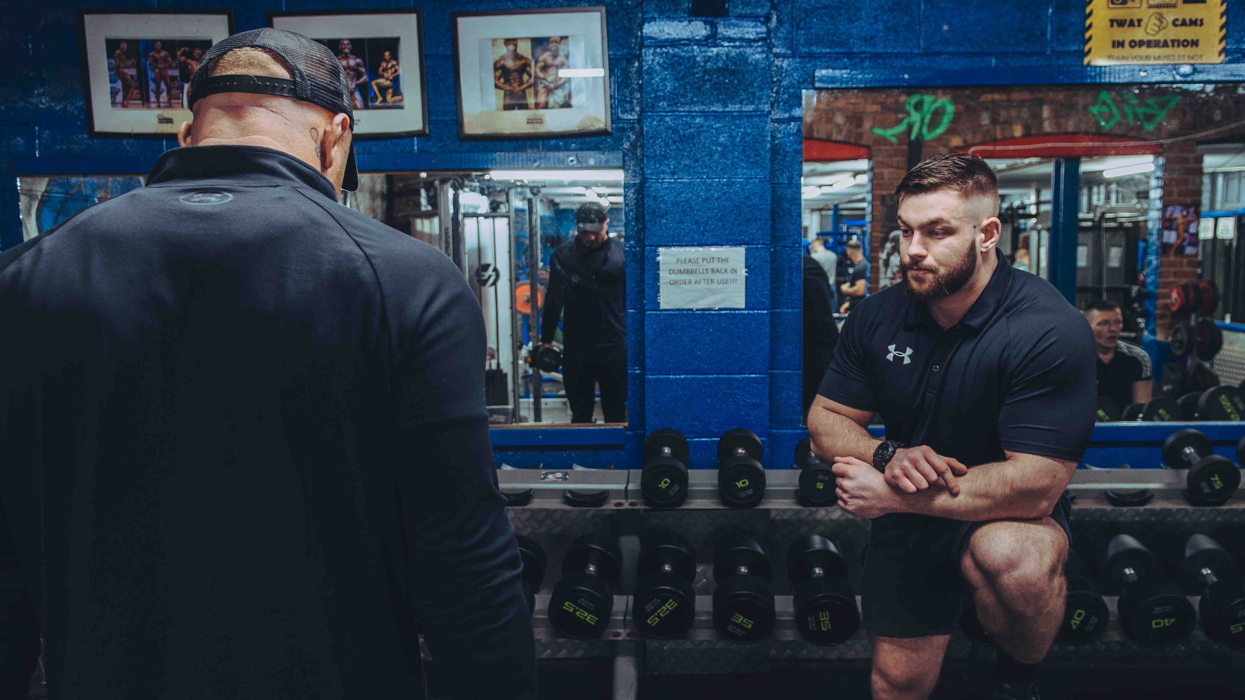 Two men in a gym with one man wearing a black cap looking at mirrors reflecting gym equipment and signs. Dumbbells are organized on racks below the mirrors. Blue gym walls with motivational signs and photos on display.