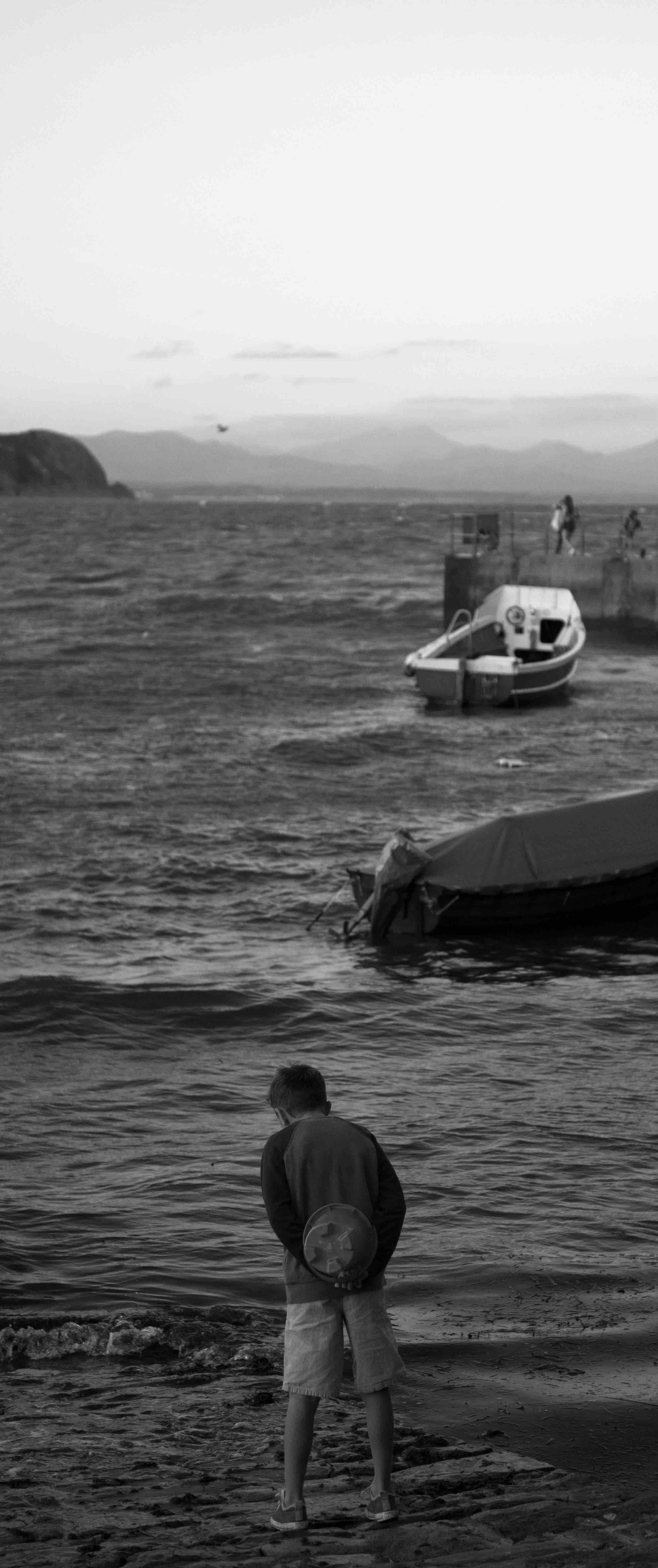Black and white photo of a person standing on a beach with boats in the water and a pier in the background.