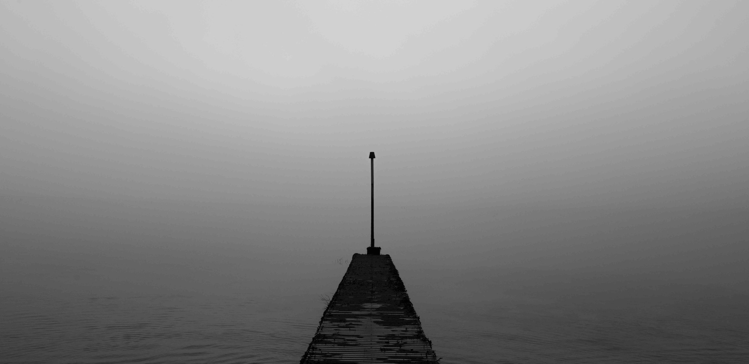 Foggy scene with a pier extending into the water and a single pole at the end in grayscale.