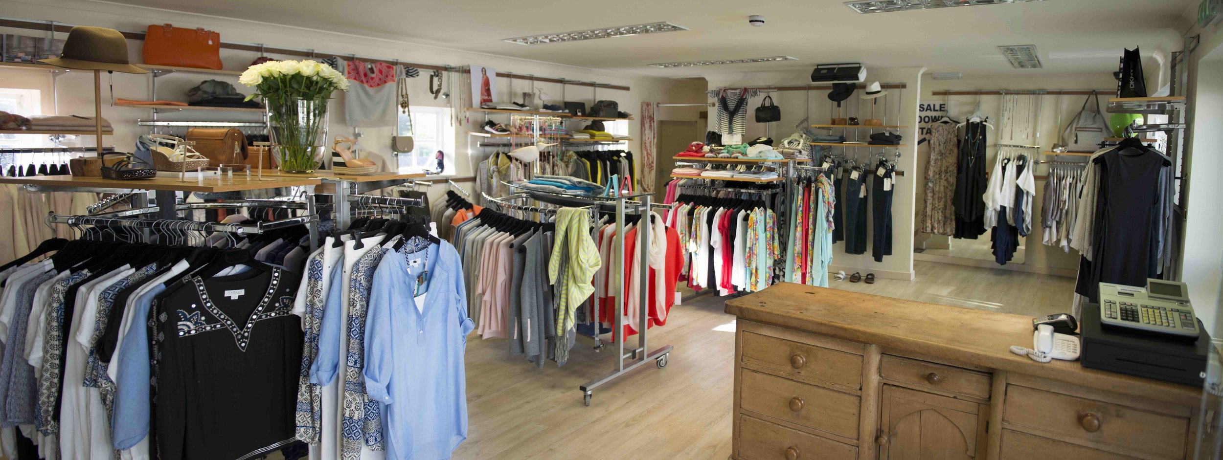 Clothing boutique with racks of colorful clothes, hats, and accessories. Display table with a vase of white flowers. Wooden counter with cash register and phone.