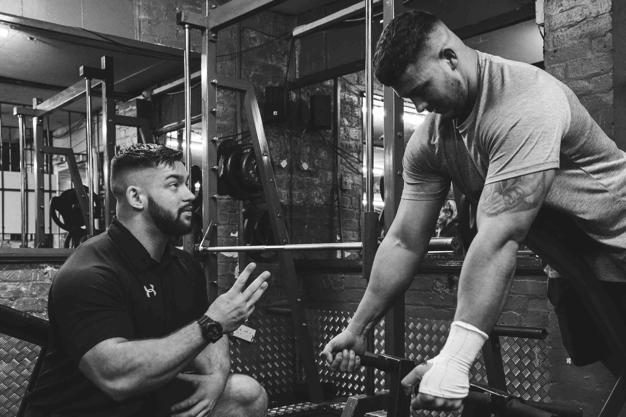 Two men in a gym, one kneeling and talking to the other who is using a workout machine. The man using the machine has a tattoo and a bandaged hand.