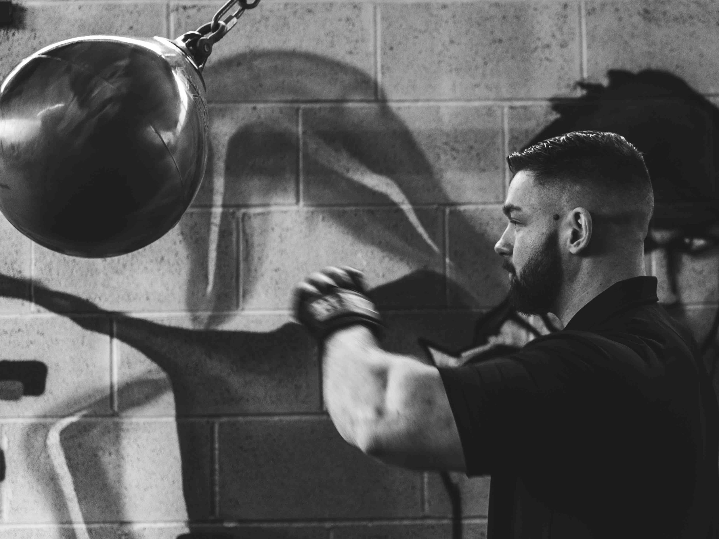 Black and white photo of a man with a beard and gloves punching a speed bag in a gym.