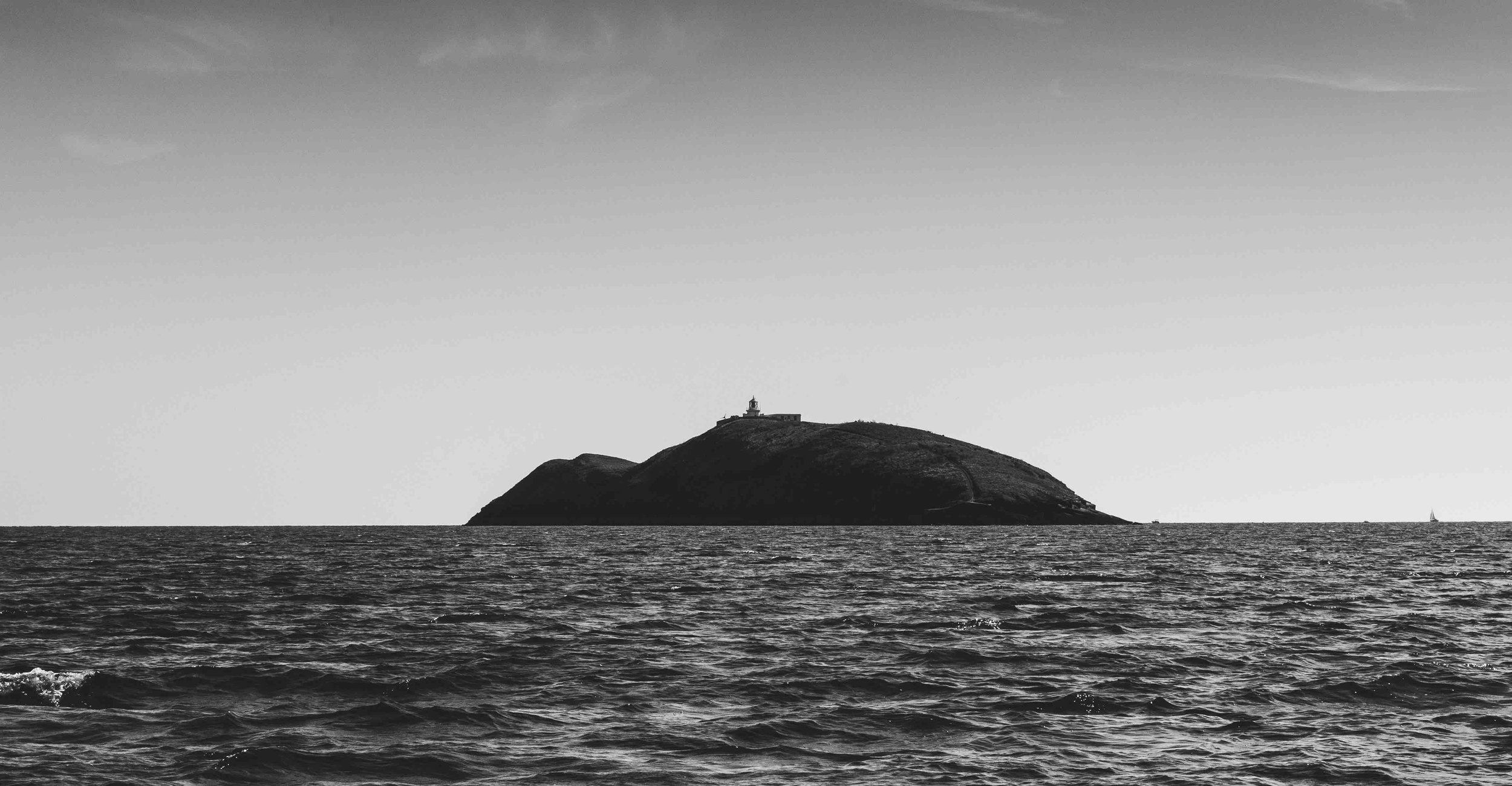 Black and white photo of an island with a lighthouse on top, surrounded by the sea.